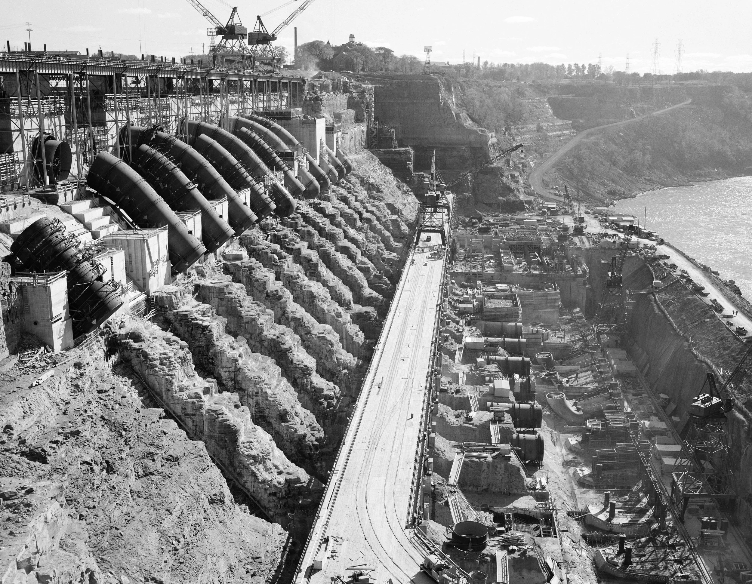 Black and white photo of a dam construction site with large pipes, cranes, and scaffolding along a rocky cliffside by a river.