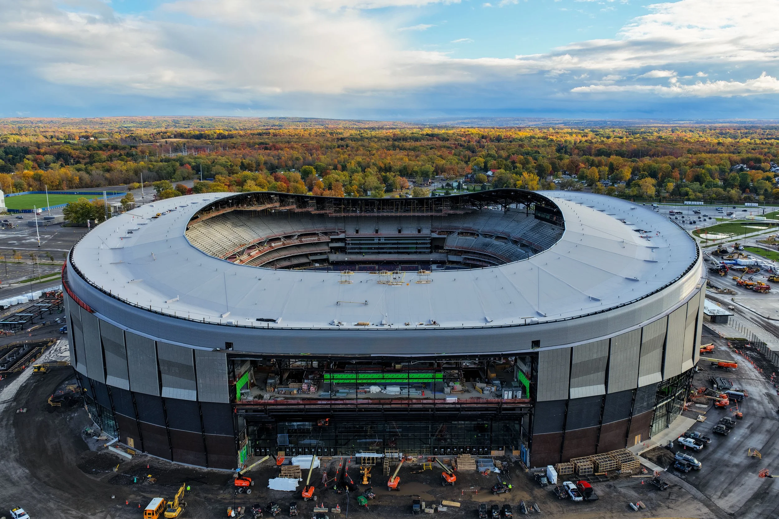 An aerial view of a large, modern stadium under construction, surrounded by trees and parking lots, with a cloudy sky overhead.