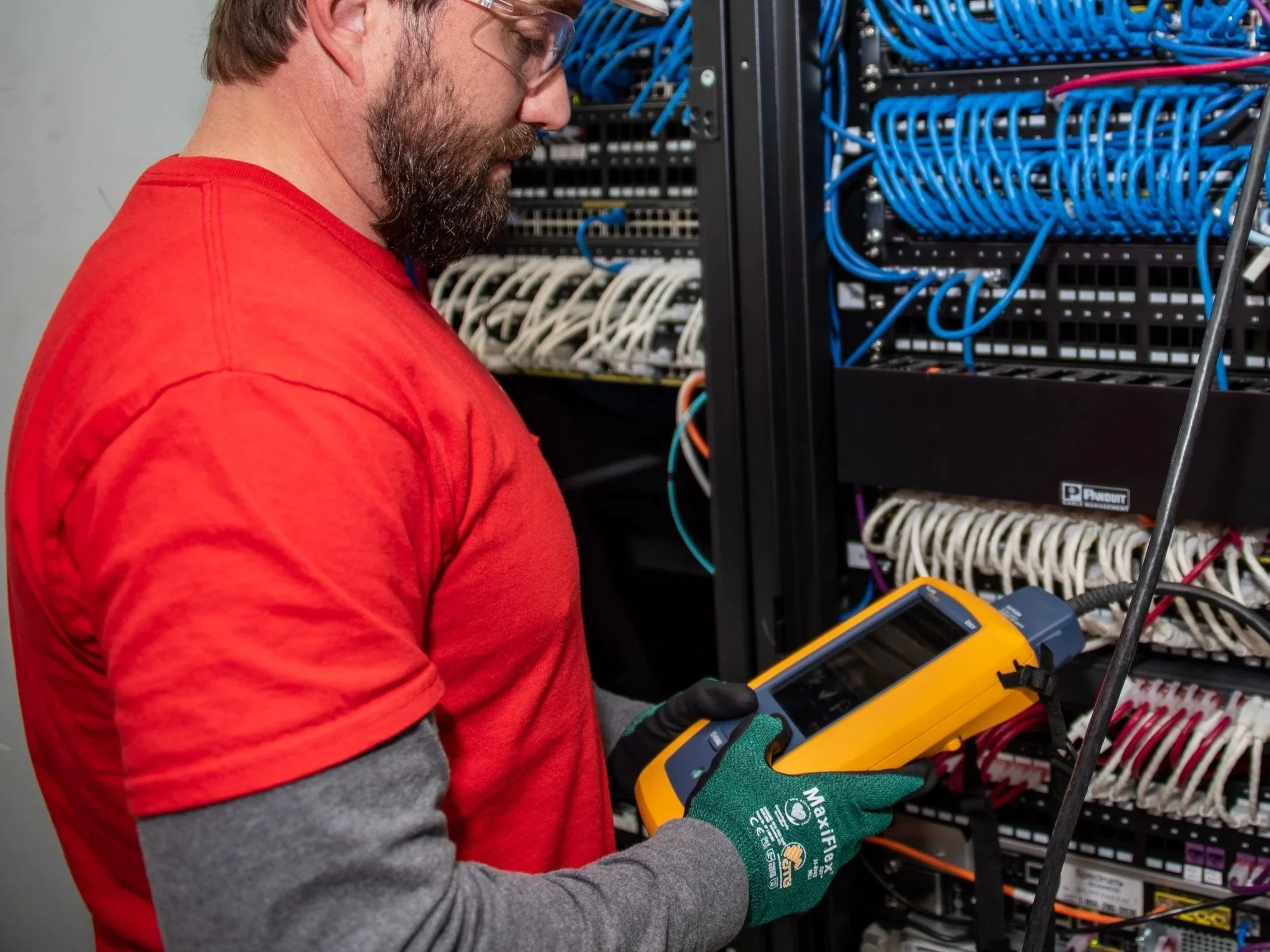 A technician wearing safety glasses, a red shirt, and green gloves is working on a server rack filled with blue and white cables in a data center.