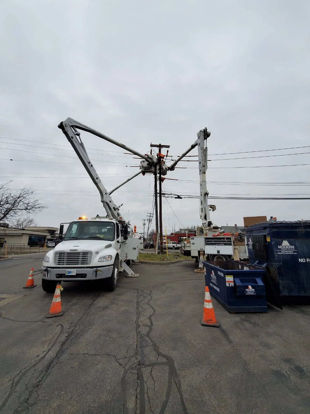Utility trucks with cherry pickers working on power lines on a cloudy day in an urban area, with orange safety cones and trash bins in the foreground.