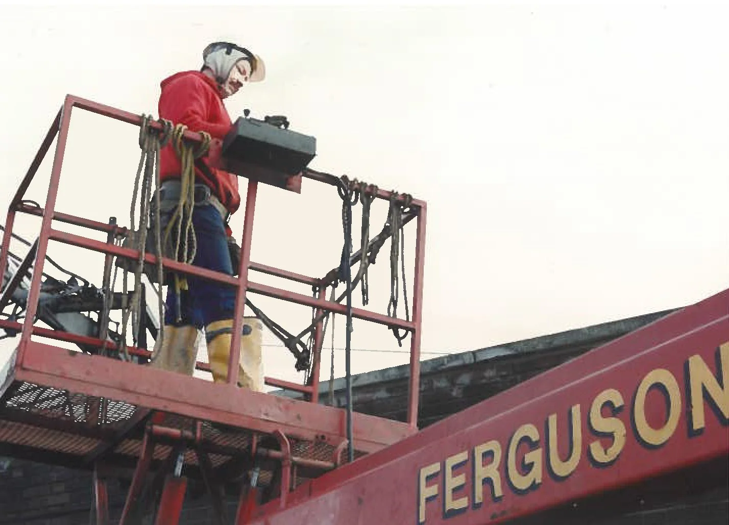 A worker in safety gear on a hydraulic lift, working on utility or street lighting installation, with the part of a sign reading 'FERGUSON' visible.
