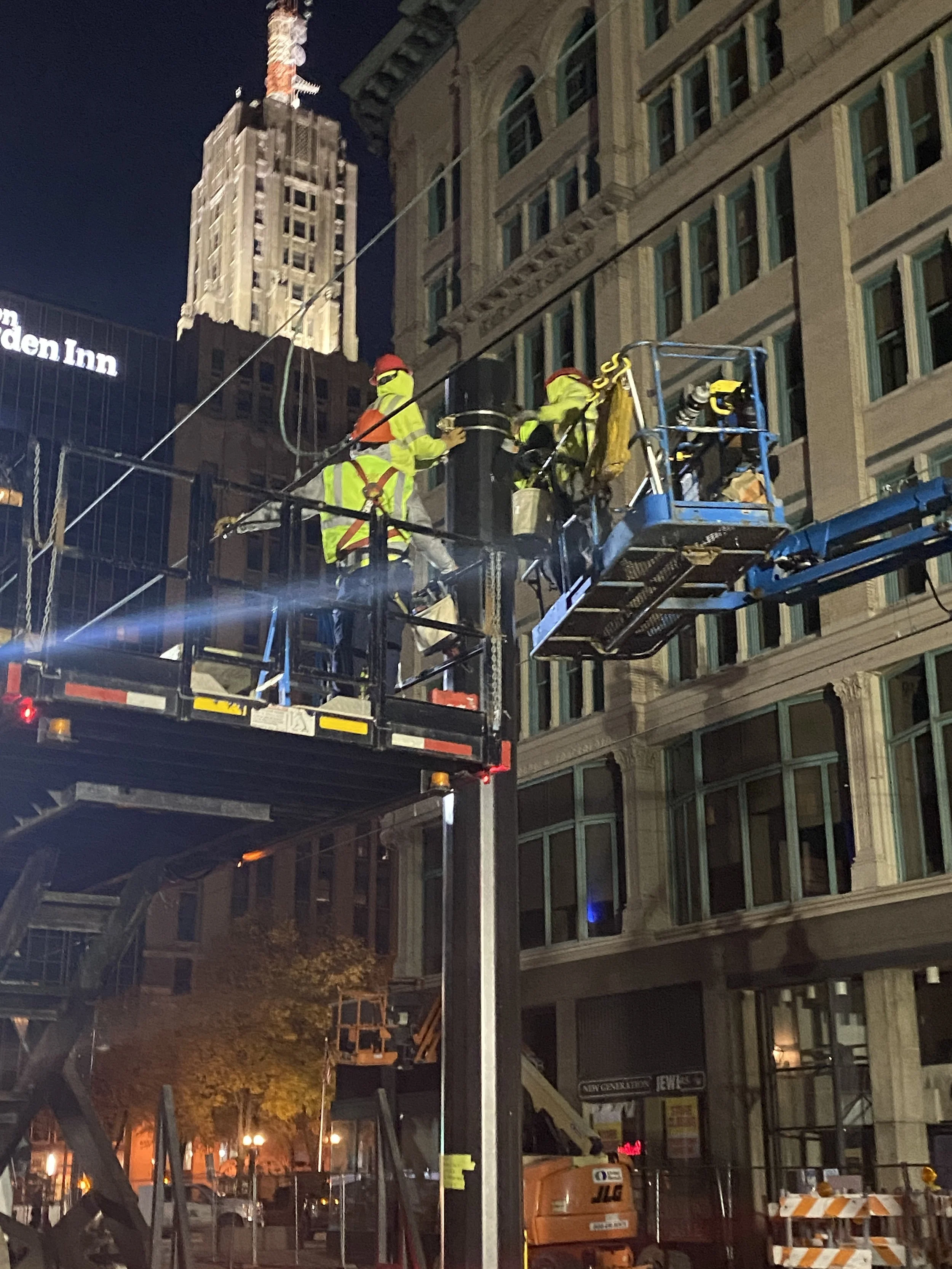 Nighttime construction scene with workers in high-visibility clothing and helmets working on an elevated platform outside a multi-story building with classic architectural details. City buildings and trees are visible in the background.