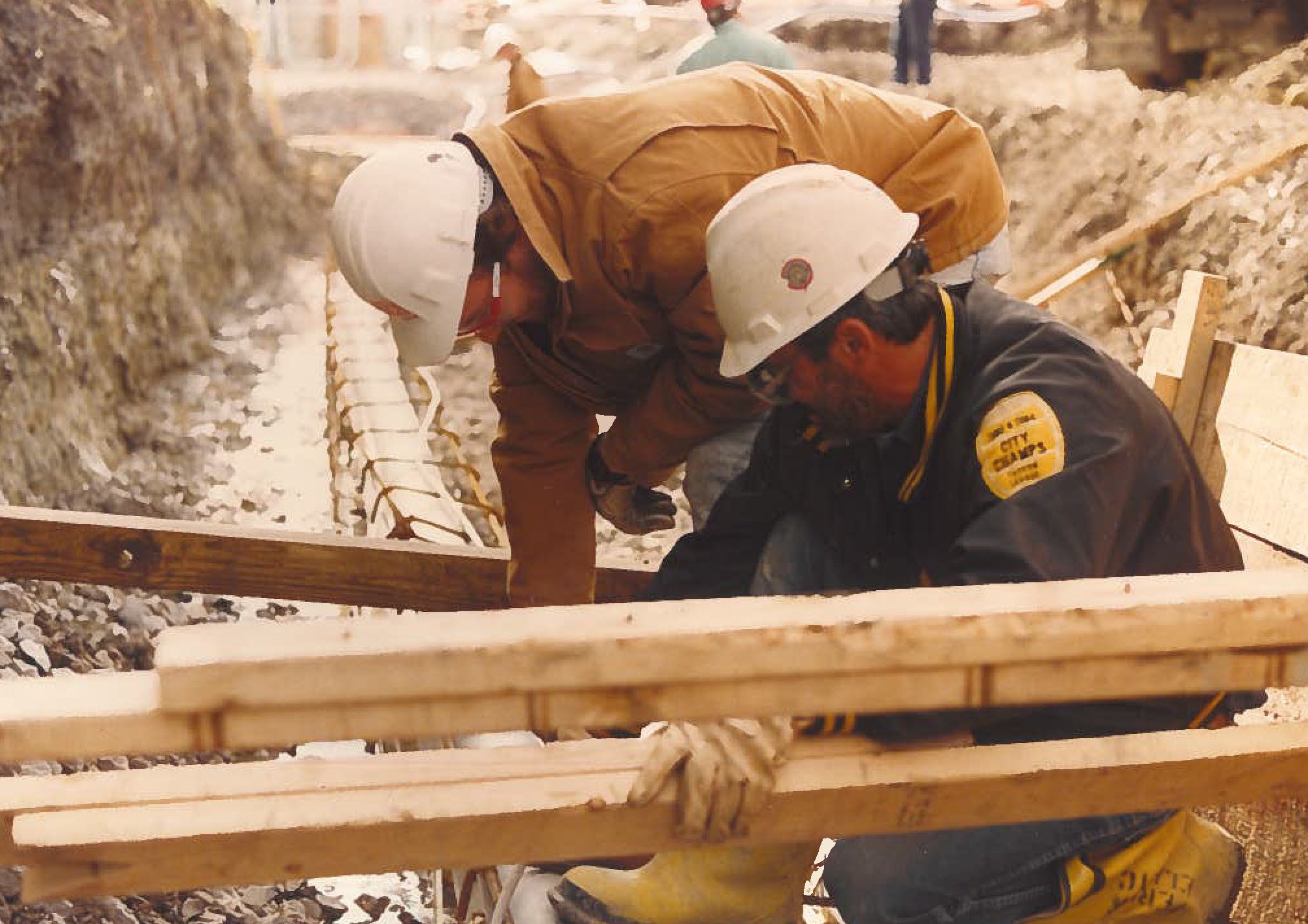 Construction workers wearing helmets and safety gear working on a building foundation, measuring and aligning wooden formwork for concrete pouring.