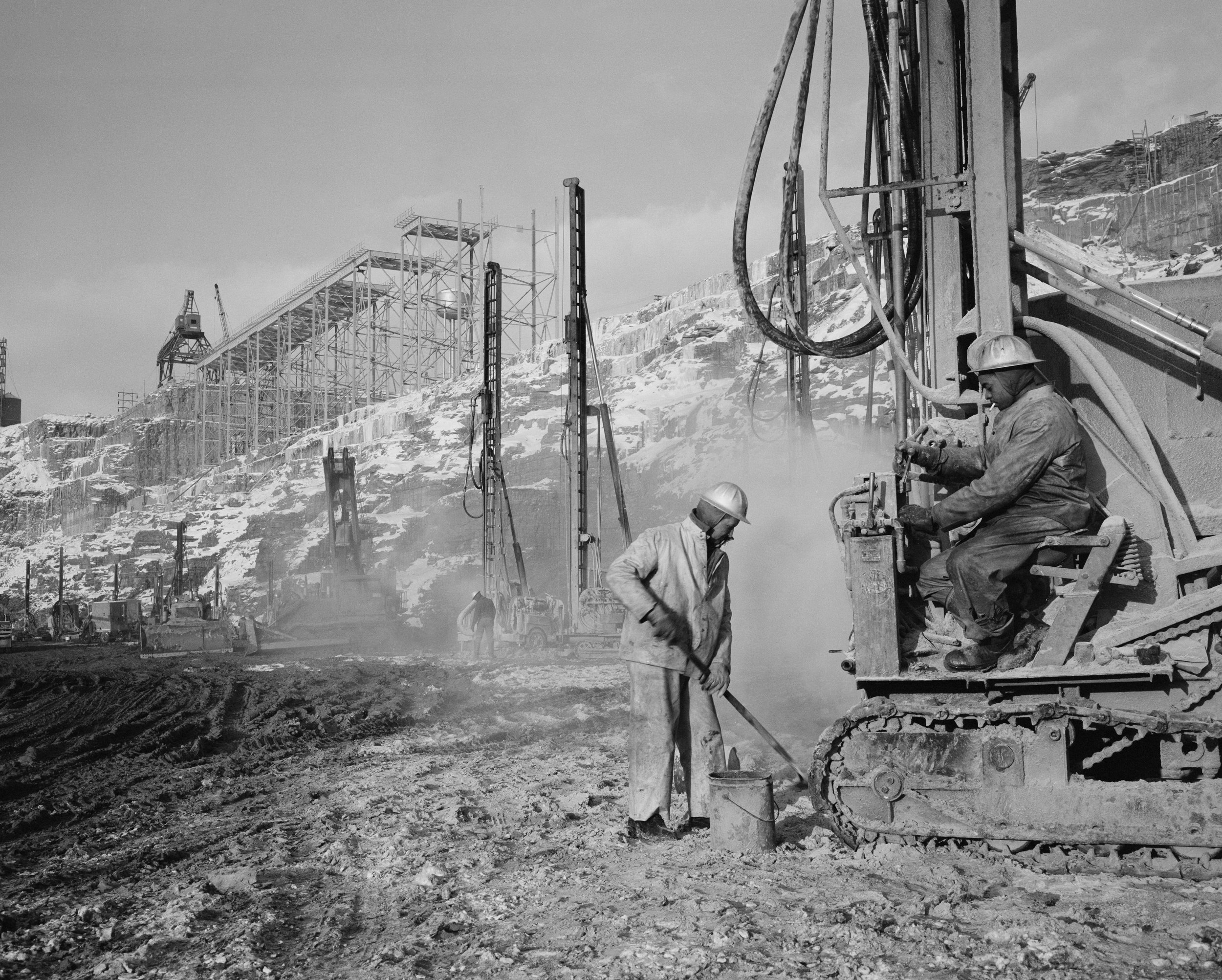 Construction workers wearing helmets working at a construction site with heavy machinery in a snowy mountainous area.
