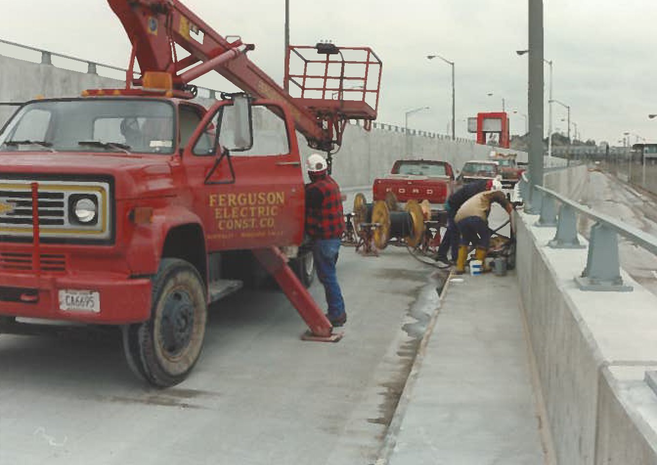 Workers repairing the sidewalk next to a highway, with a red Ferguson Electric utility truck and a Ford truck in the background.