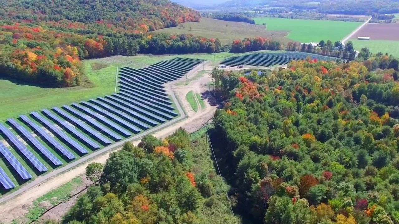 Aerial view of solar panels on a sloped hillside surrounded by a forest with autumn foliage, farmland, and open fields in the background.