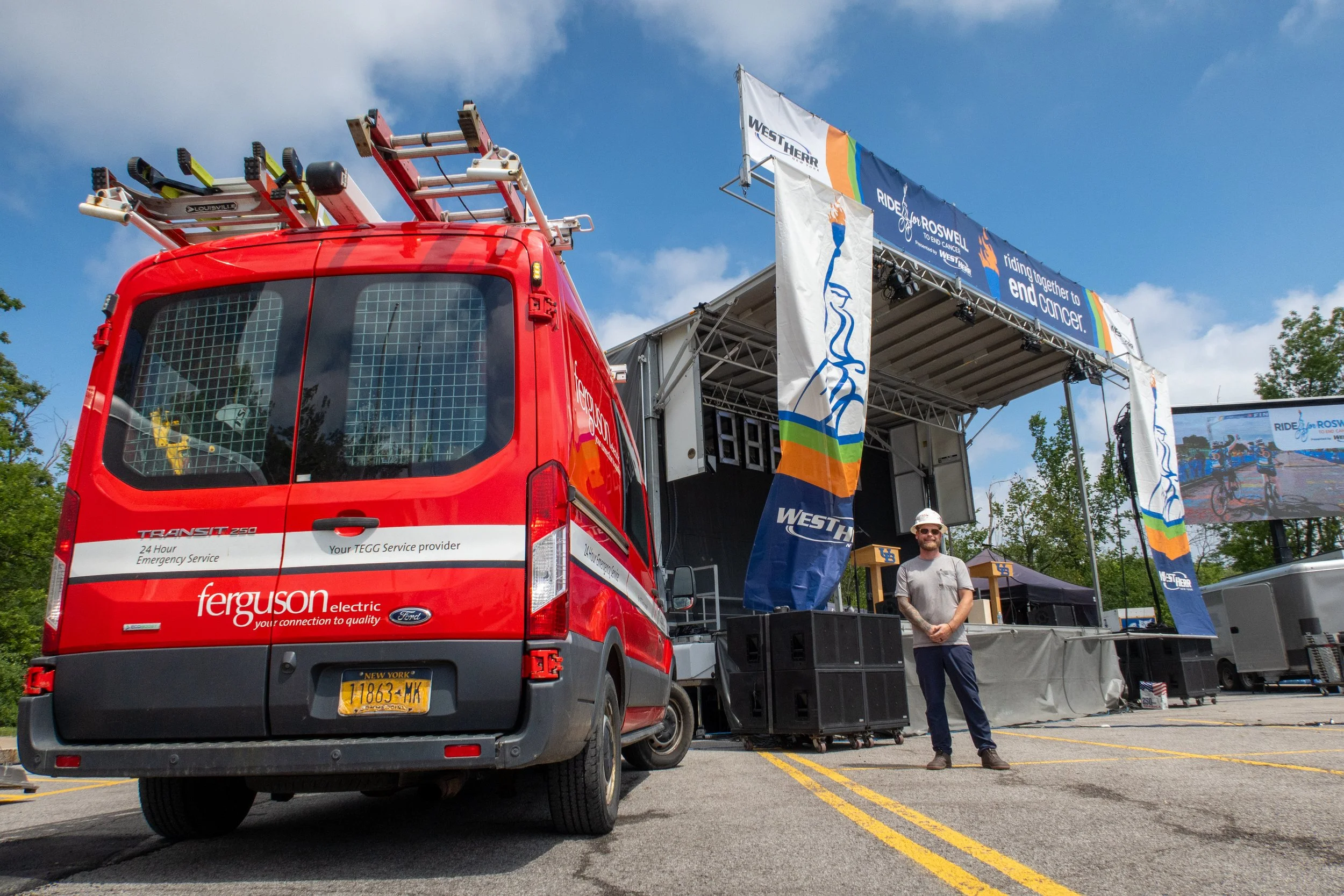 A red Ferguson utility van with emergency lights parked in a parking lot, with a large stage in the background displaying banners for the Ride for Roswell event and a man standing nearby. The stage has a banner that reads 'riding together to end cancer' and various flags and banners. The sky is partly cloudy.