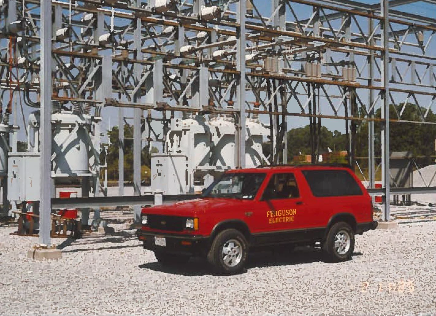 A red electric utility vehicle marked 'Ferguson Electric' parked in front of electrical infrastructure, including transformers and metal framework.