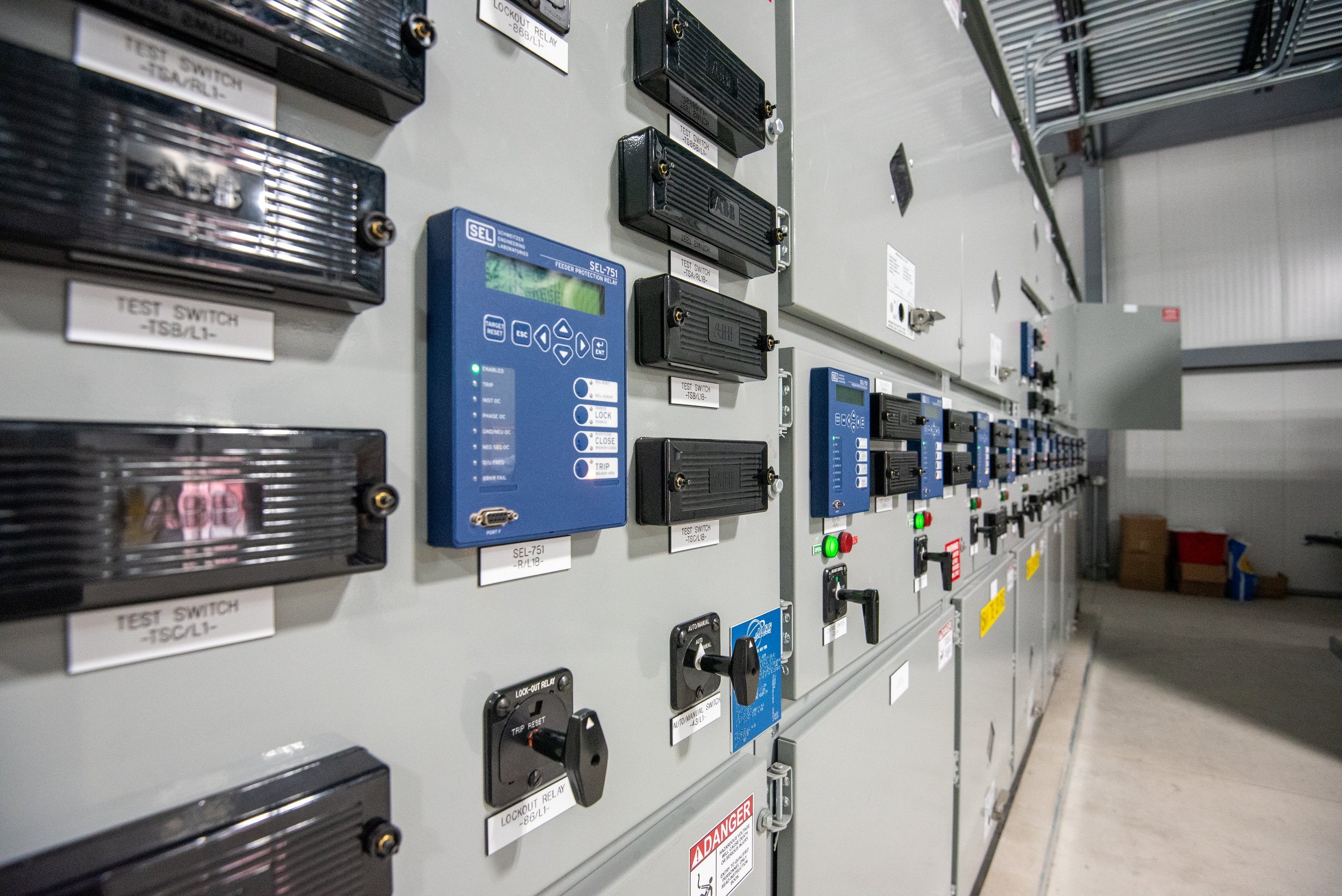 An electrical control panel with multiple switches, relays, and circuit breakers, labeled with various tags and some with indicator lights, located in an industrial or utility setting.