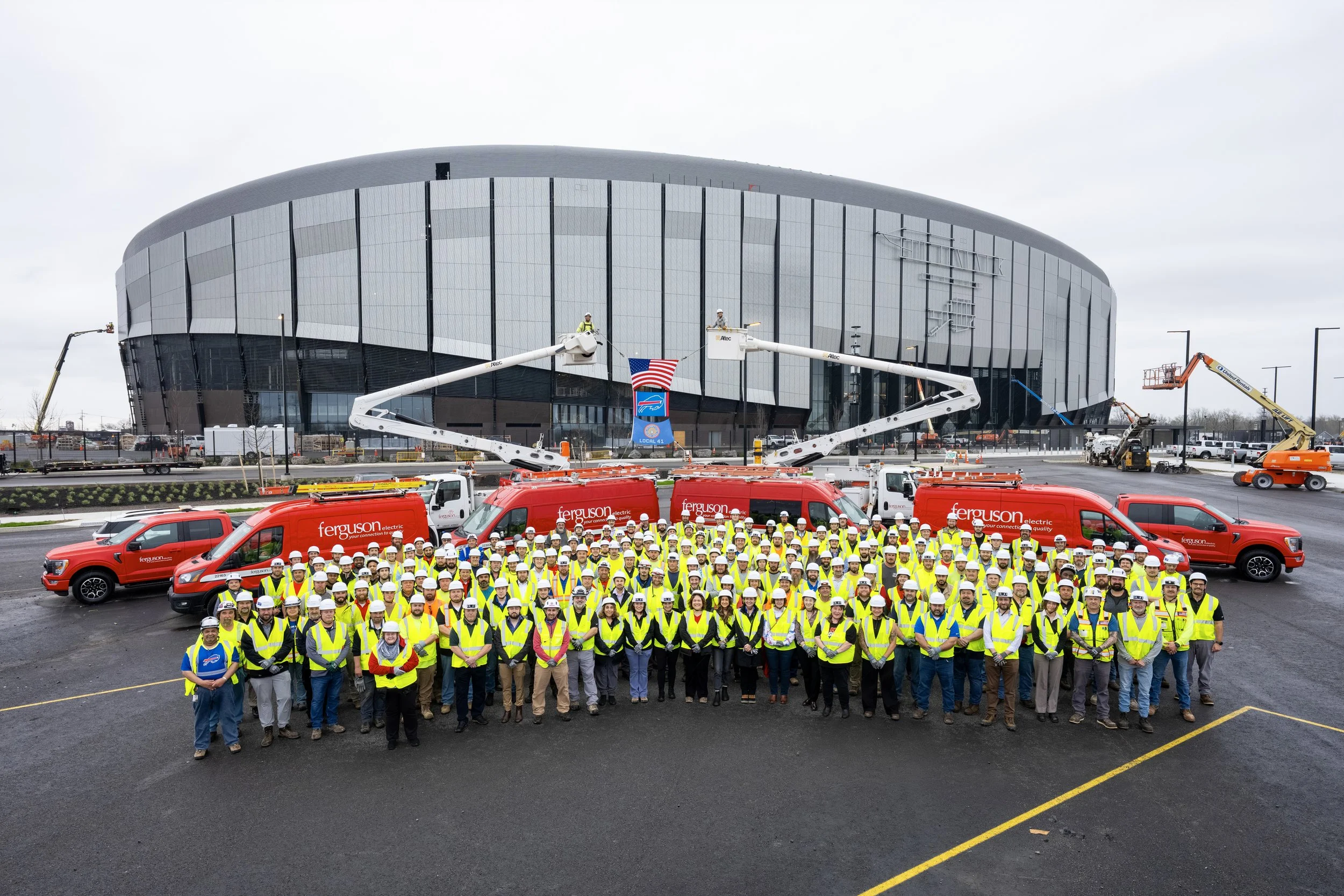 Group of construction workers and staff in yellow safety vests and helmets standing in front of a stadium during daytime