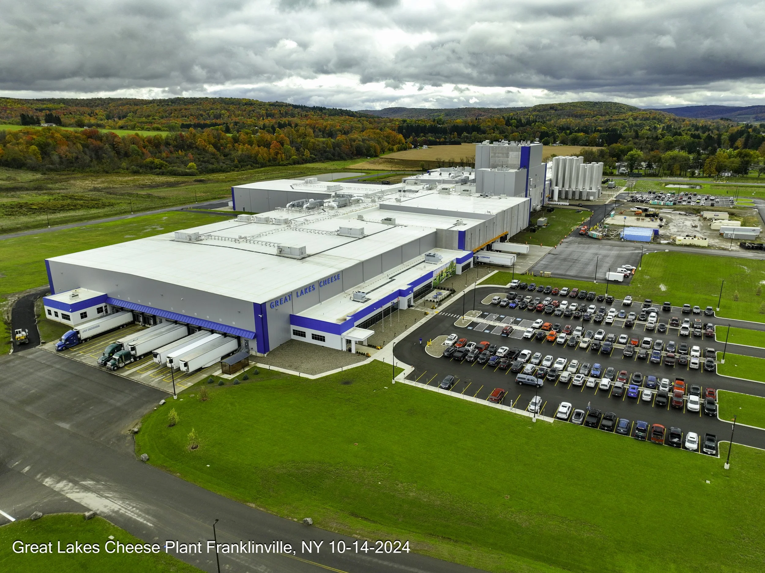 Aerial view of Great Lakes Cheese plant in Franklinville, NY, with large white industrial buildings, parking lot filled with cars, and surrounding green fields and forested hills under cloudy sky.