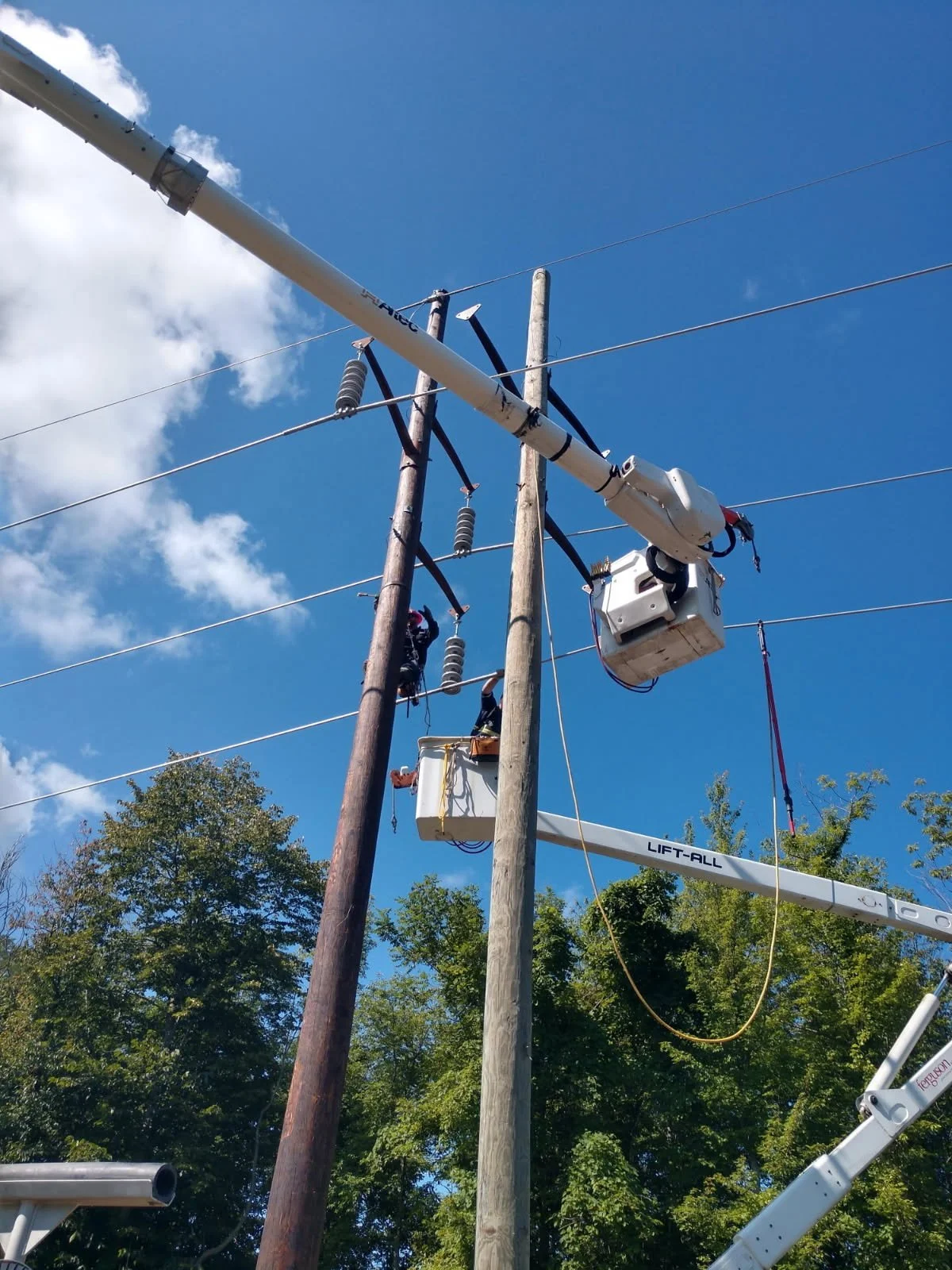Workers on cherry pickers repairing electrical poles and wires outdoors under a bright blue sky with clouds.