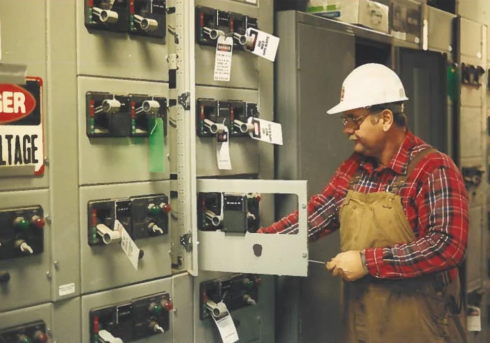 A worker in a hard hat and plaid shirt working on electrical control panels in an industrial setting.