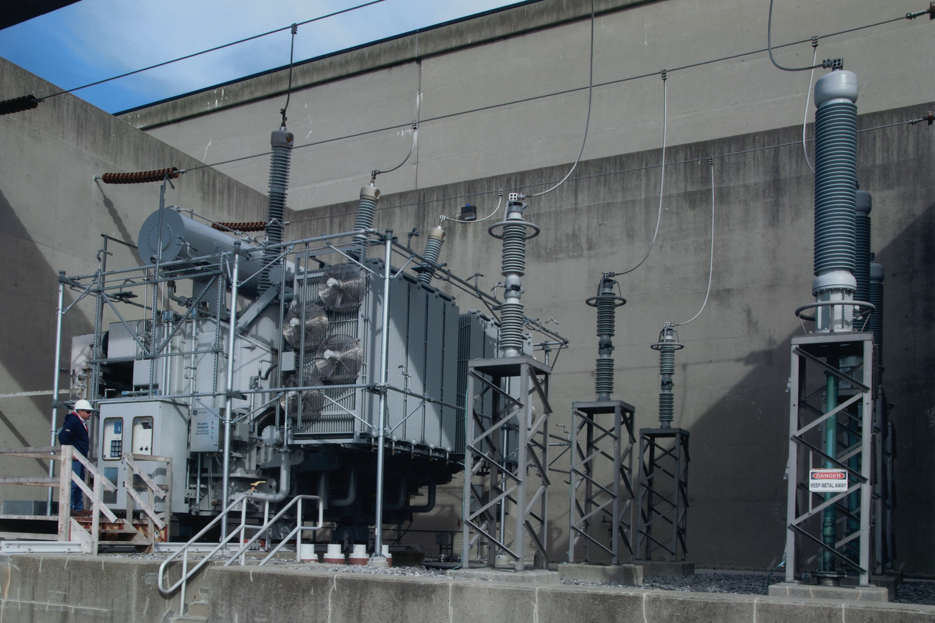 Industrial electrical equipment and transformers installed outdoors against a concrete wall, with safety signage and a worker in a white helmet.