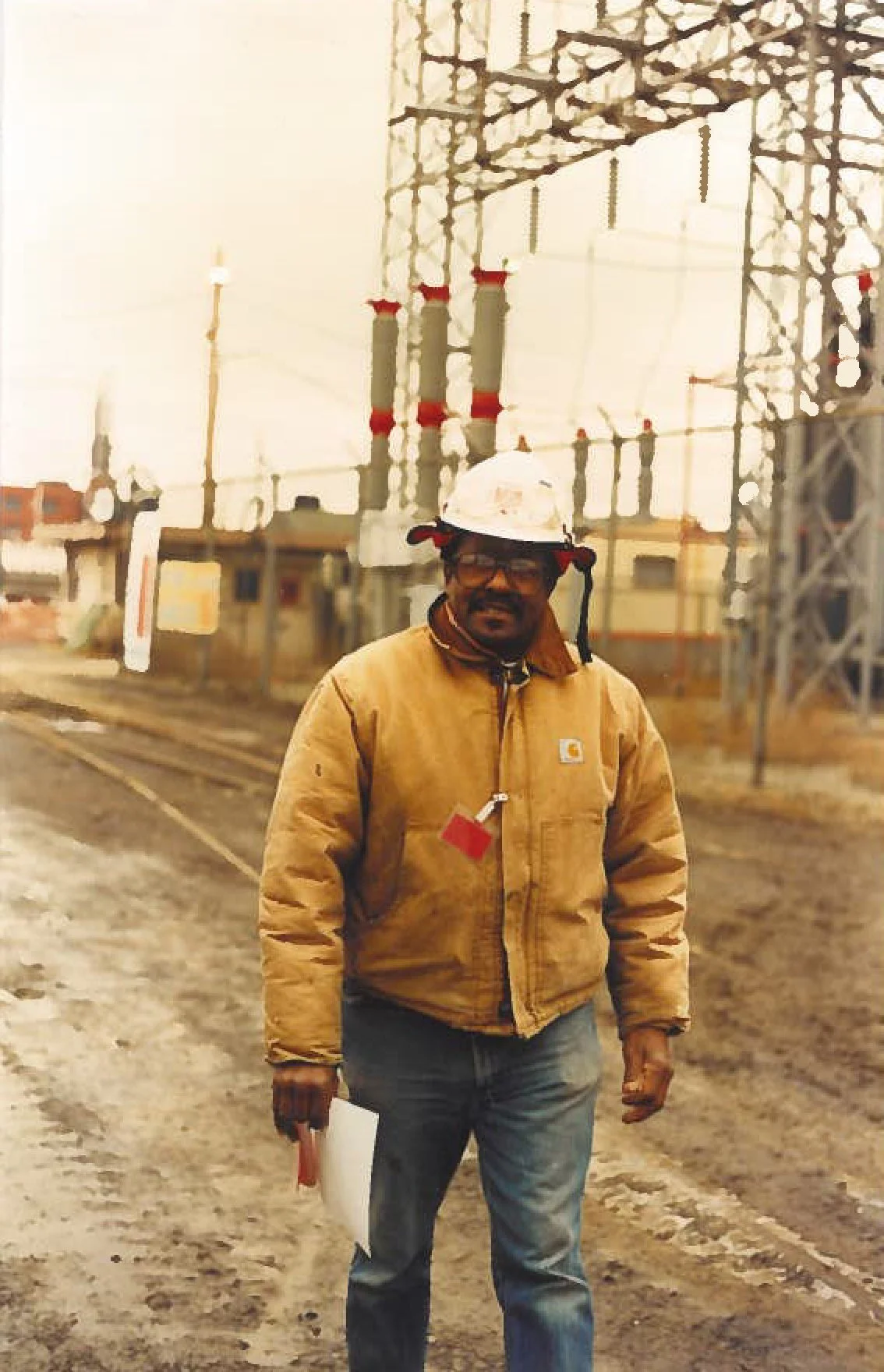 A man standing outdoors near industrial power station equipment, wearing a yellow jacket, glasses, and a white hard hat with a red clip, holding a rolled paper in his right hand.