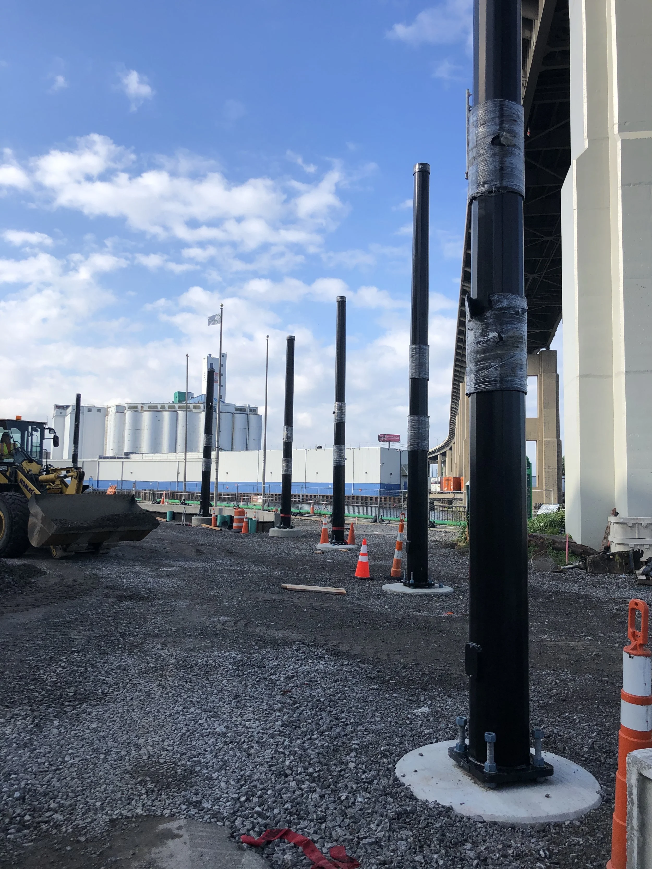 Construction site with black metal poles installed on concrete bases, orange safety cones, construction equipment, and a bridge with support beams in the background under a partly cloudy sky.