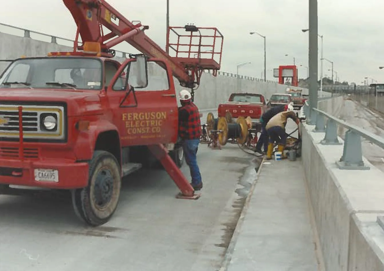 Workers repairing a concrete barrier on a highway with a red Ferguson electric utility truck and a work lift, under an overcast sky.