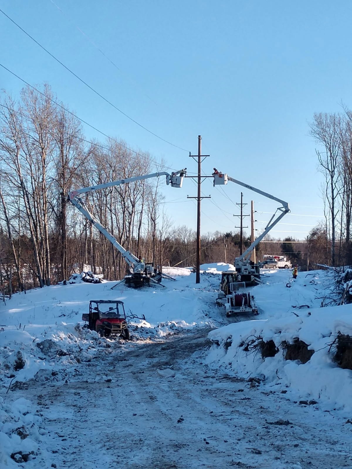 Workers using bucket trucks to repair power lines on a snowy landscape, with utility vehicles parked nearby and leafless trees in the background.