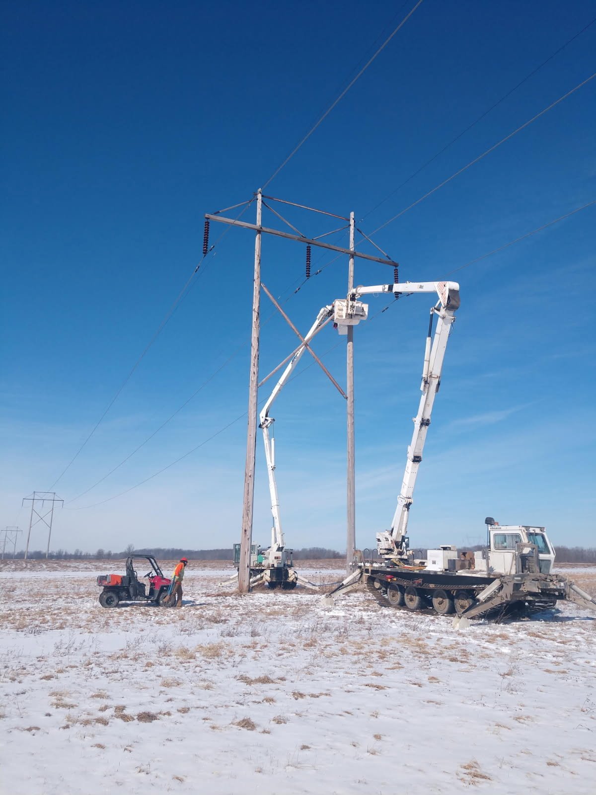 A utility worker in a hard hat operating a bucket truck on a snow-covered open field, repairing a wooden power line pole while another worker on a utility vehicle observes.