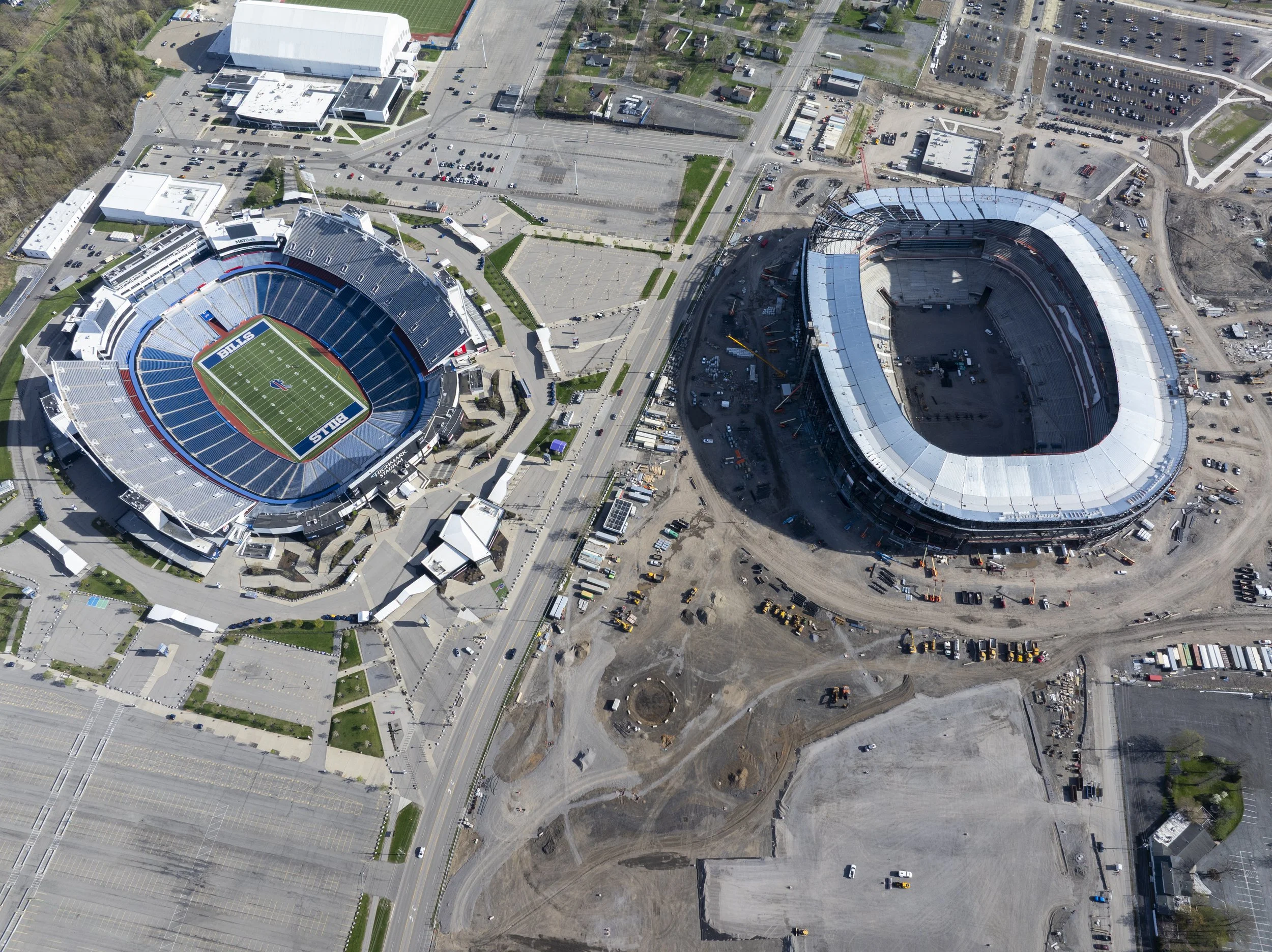 Aerial view of a stadium under construction next to a completed stadium and parking lot, with surrounding buildings and streets nearby.