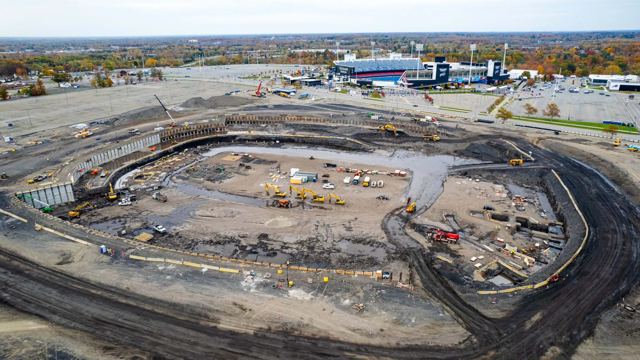 Aerial view of a large construction site with heavy machinery, dirt, and water, adjacent to a football stadium and parking lot during autumn.
