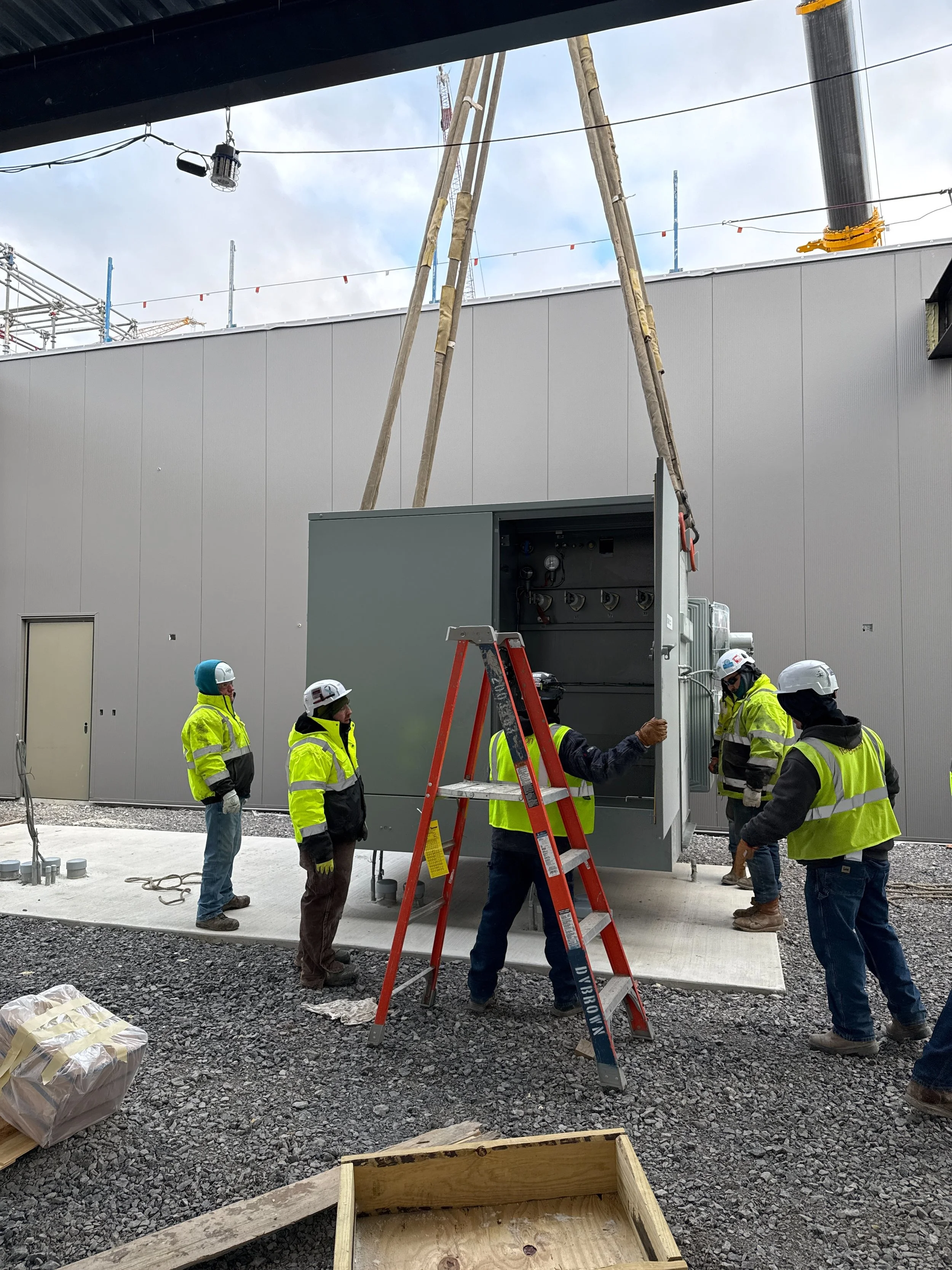 Construction workers wearing safety gear working outdoors on electrical equipment near a building under construction.