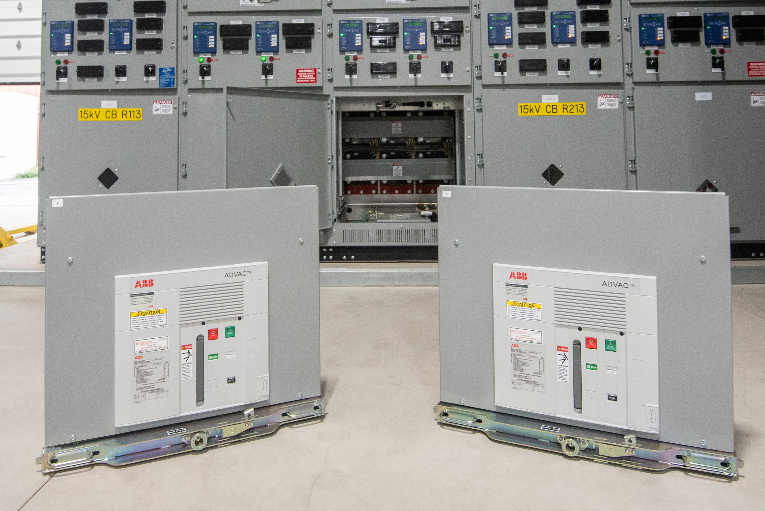 Two electrical panels on a concrete floor, with open doors revealing internal components and wiring, labeled with caution and voltage signs.