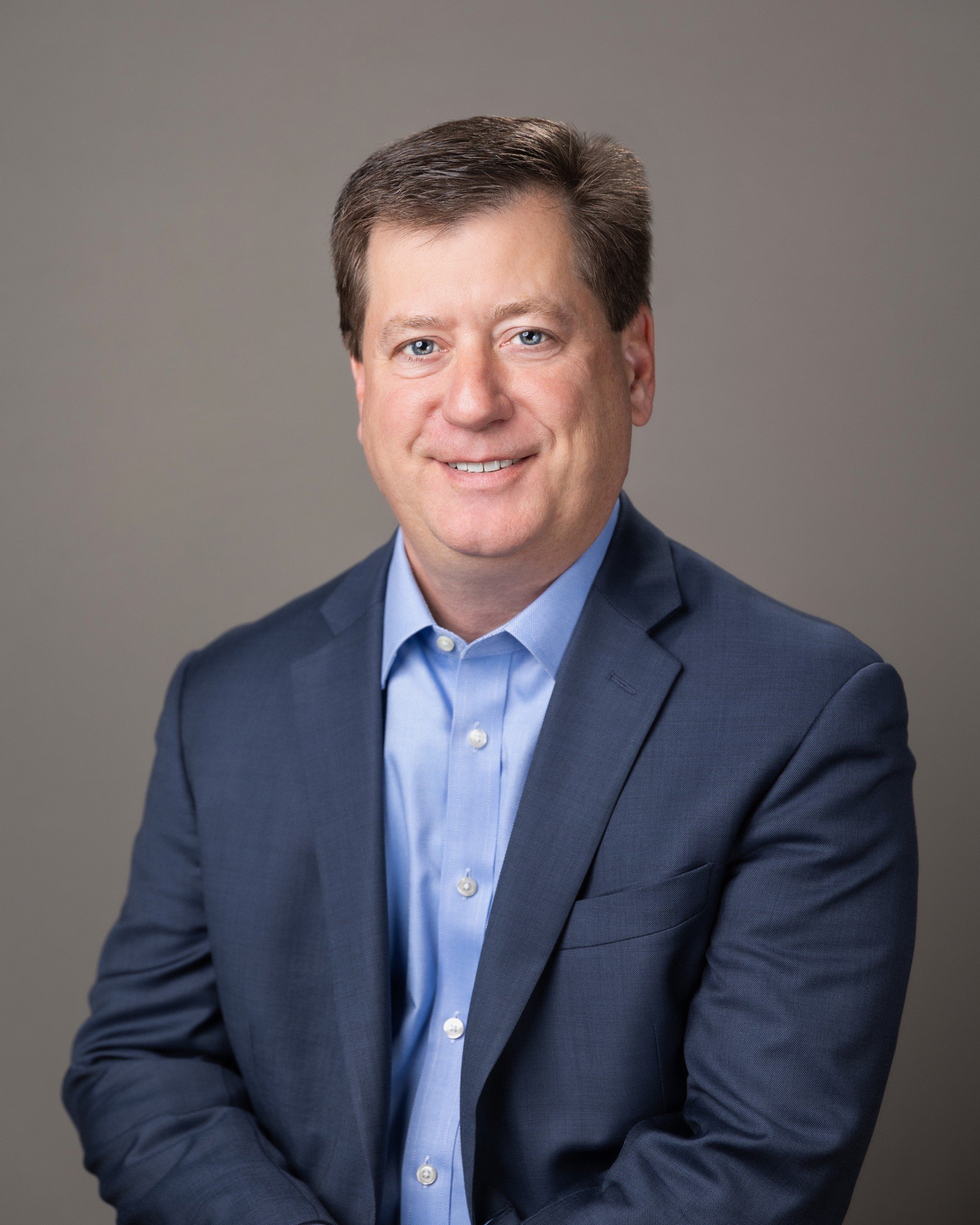 A professional man with light skin, blue eyes, dark brown hair, dressed in a navy suit and a light blue shirt, smiling at the camera against a plain gray background.