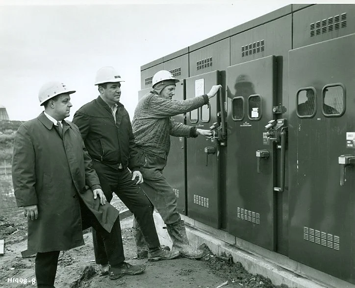 Three men in business attire and hard hats inspecting electrical or industrial equipment outdoors.