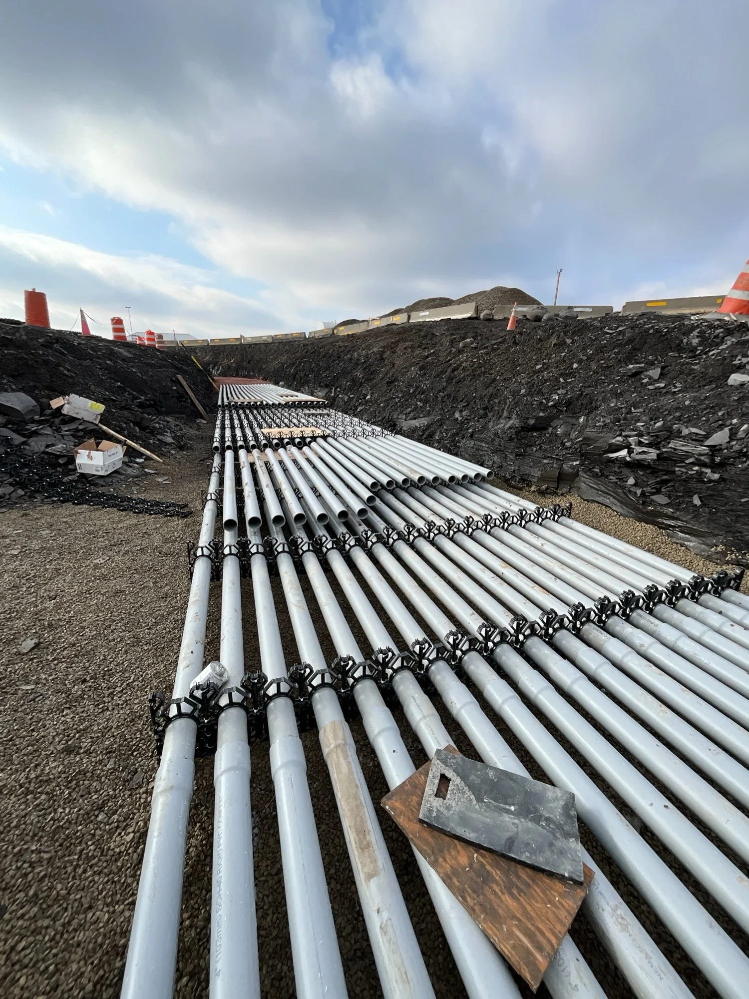 Construction site with multiple white pipes laid on gravel ground, black soil piles, orange traffic cones, and concrete barriers under cloudy sky.