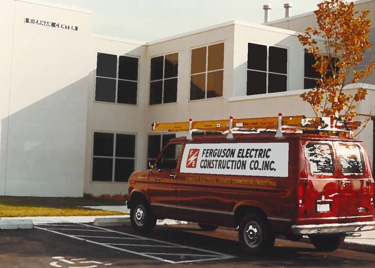 Red van with Ferguson Electric Construction Co., Inc. sign parked in front of a white building with large windows and a tree with orange leaves.