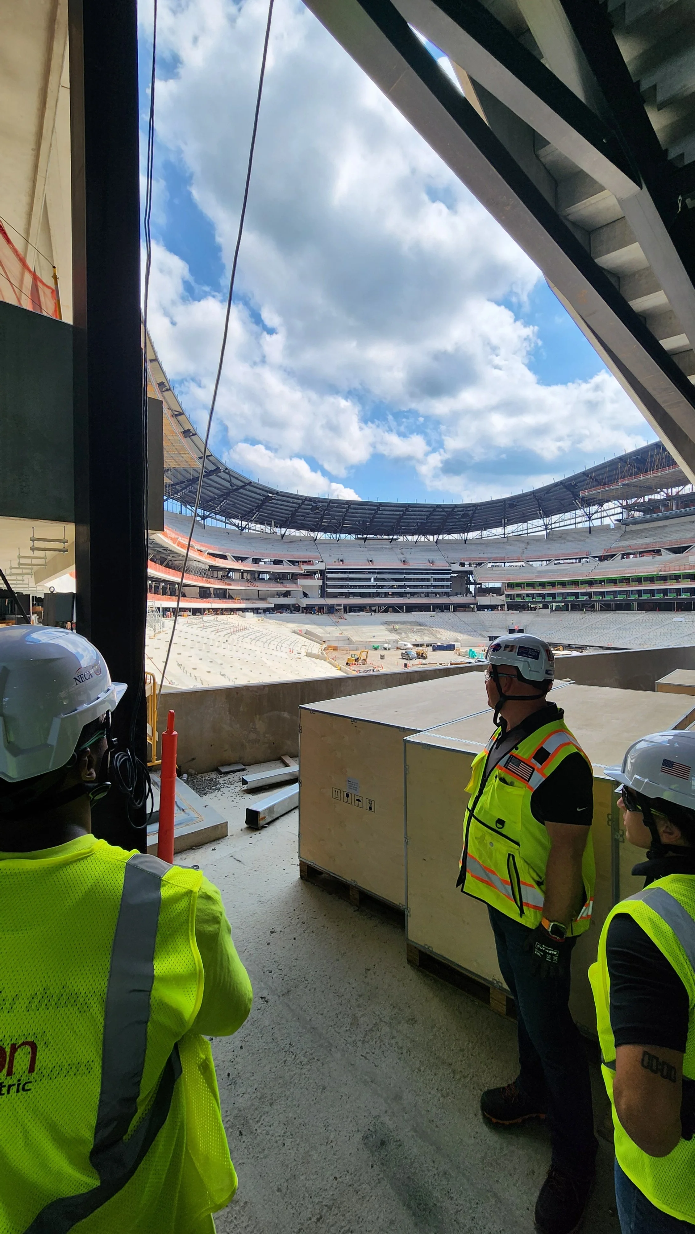 Workers in yellow safety vests and hard hats standing inside a large stadium under construction. The stadium has multiple levels of seating and a partially completed roof. The sky is partly cloudy.