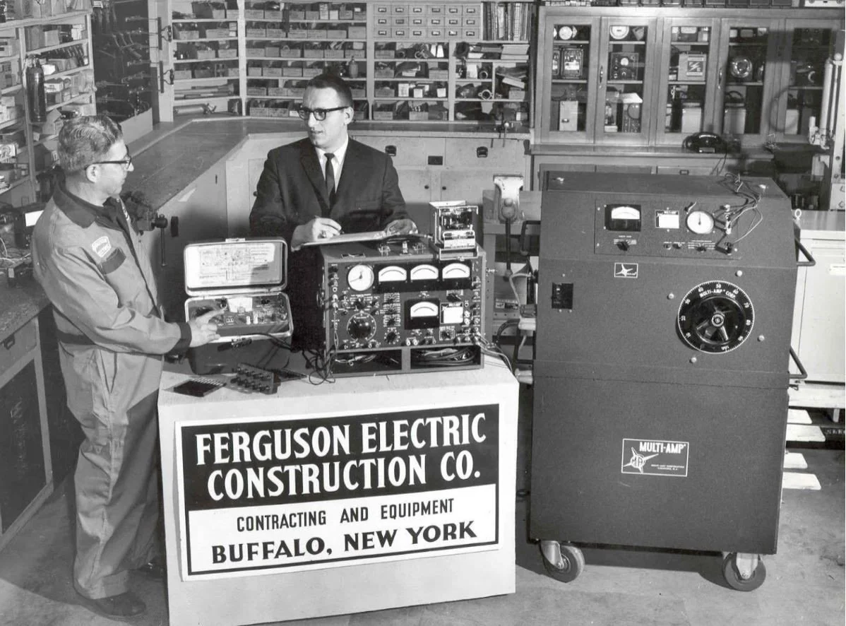 Two men are working with electrical testing equipment in a workshop. One man is holding a box-like device, and the other man is writing or adjusting a control panel. There are various machines and tools, and a sign in front reads 'Ferguson Electric Construction Co., Buffalo, New York.'