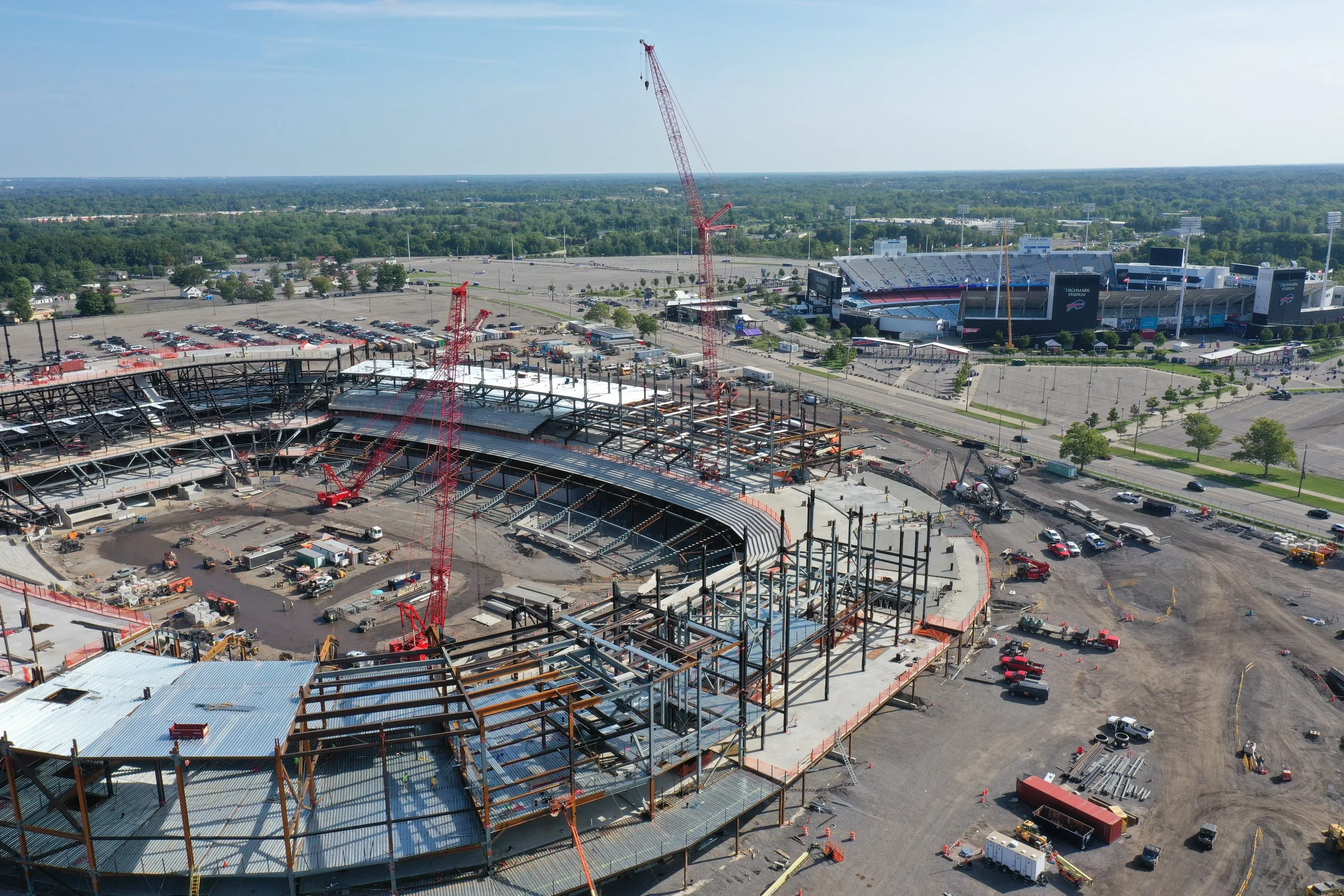 Construction site of a stadium with steel framework being assembled, several cranes, parking lots, and a nearby stadium building labeled 'Highmark Stadium' in the background.