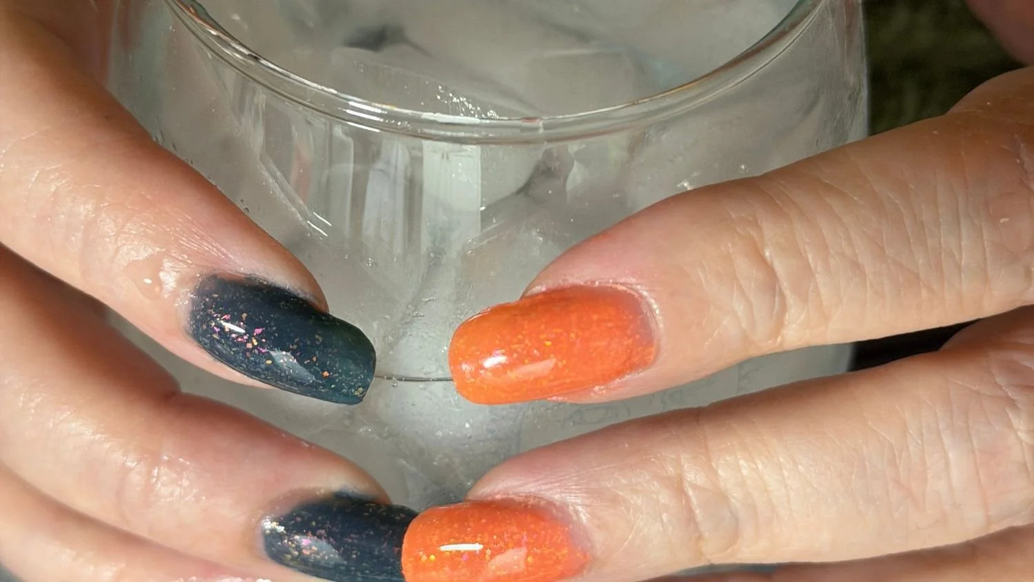 Two hands holding a condensation-covered glass of ice water, nails showing You're Soaking In It thermochromic shift in real time — cool state fingers in slate blue-gray with iridescent flake scatter, warm state fingers in vintage burnt orange.