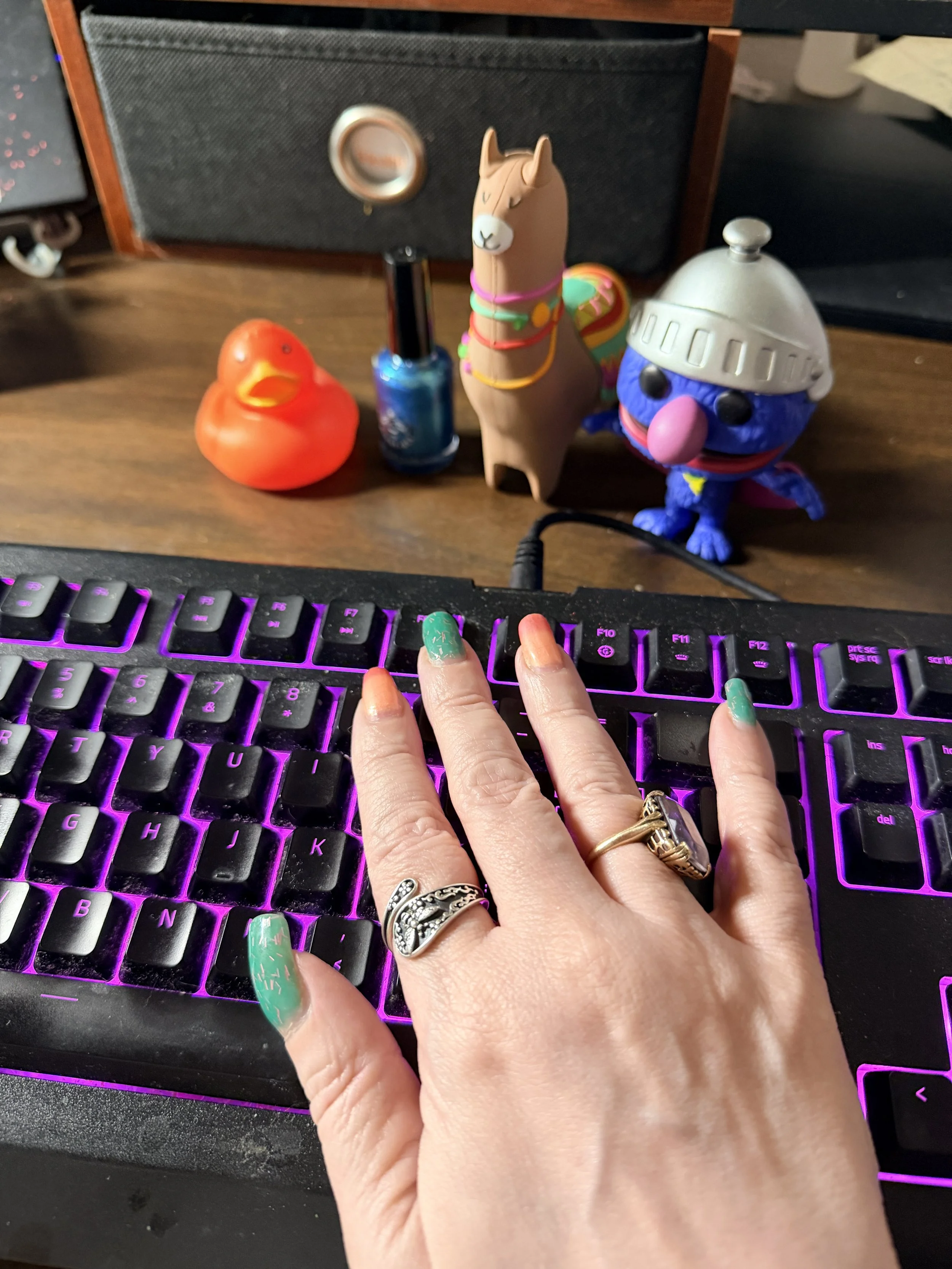 Hand with peach and teal nails totally not posed on a keyboard with duck, llama, and Super Grover bearing witness.