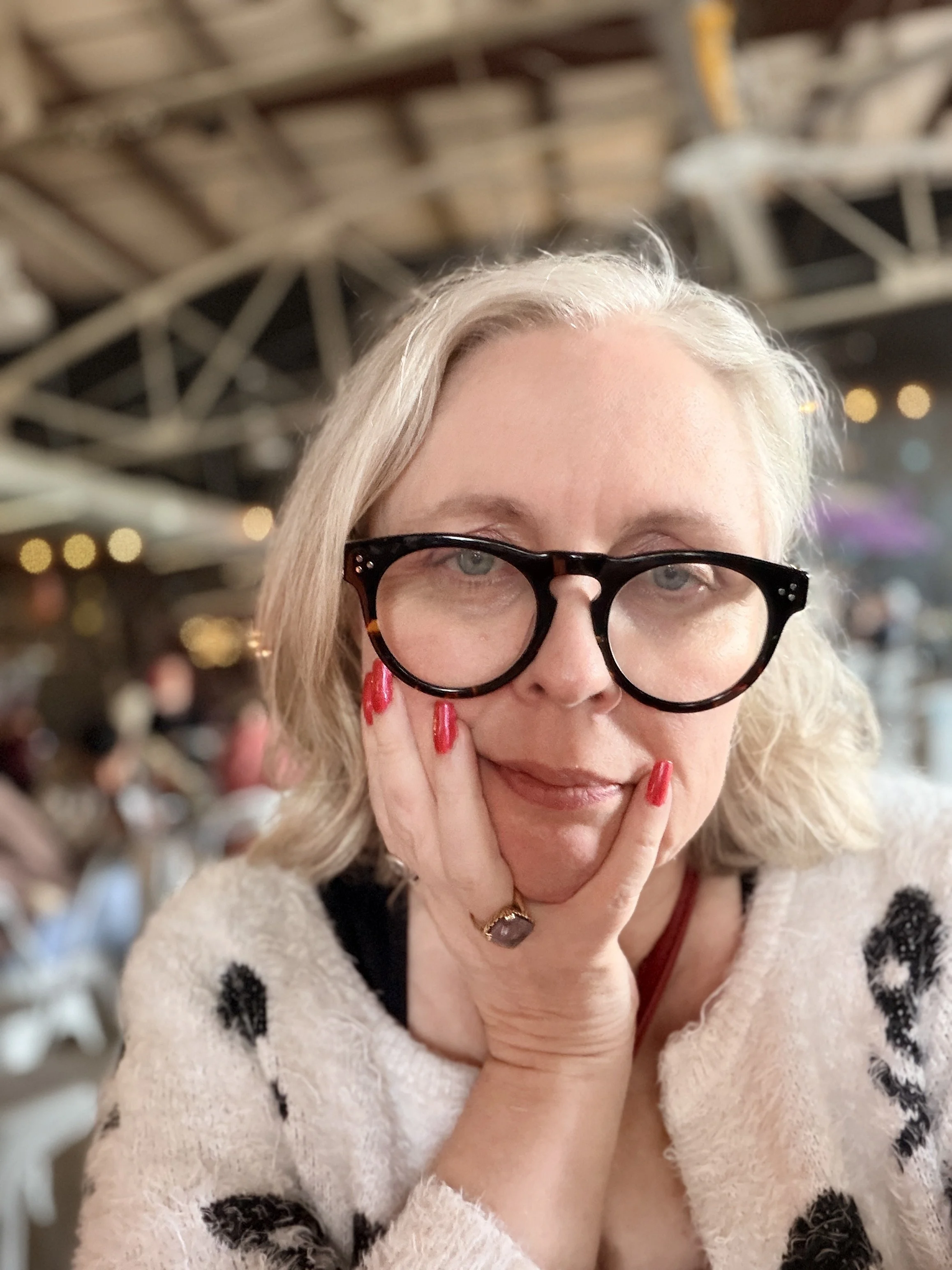 Founder Heather McGrew in a white skull-and-crossbones cardigan, deadpan expression, hand on cheek, red shimmer nails visible. Bad Astronaut in background.