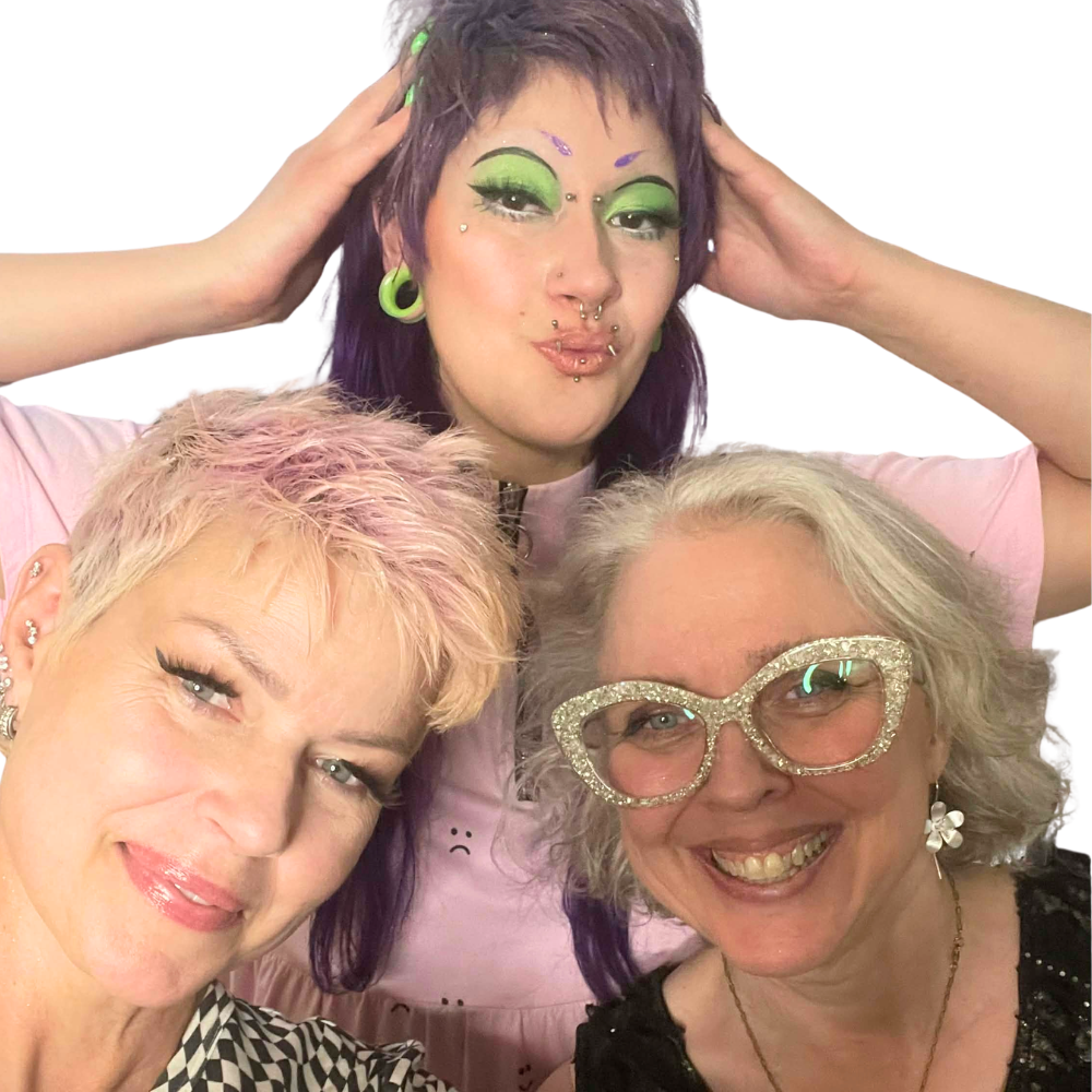 Three women of different ages and styles laugh together against a white background — pink pixie hair, purple hair with green eye makeup, and silver hair with glittery cat-eye glasses. Real people, no filter, all welcome.