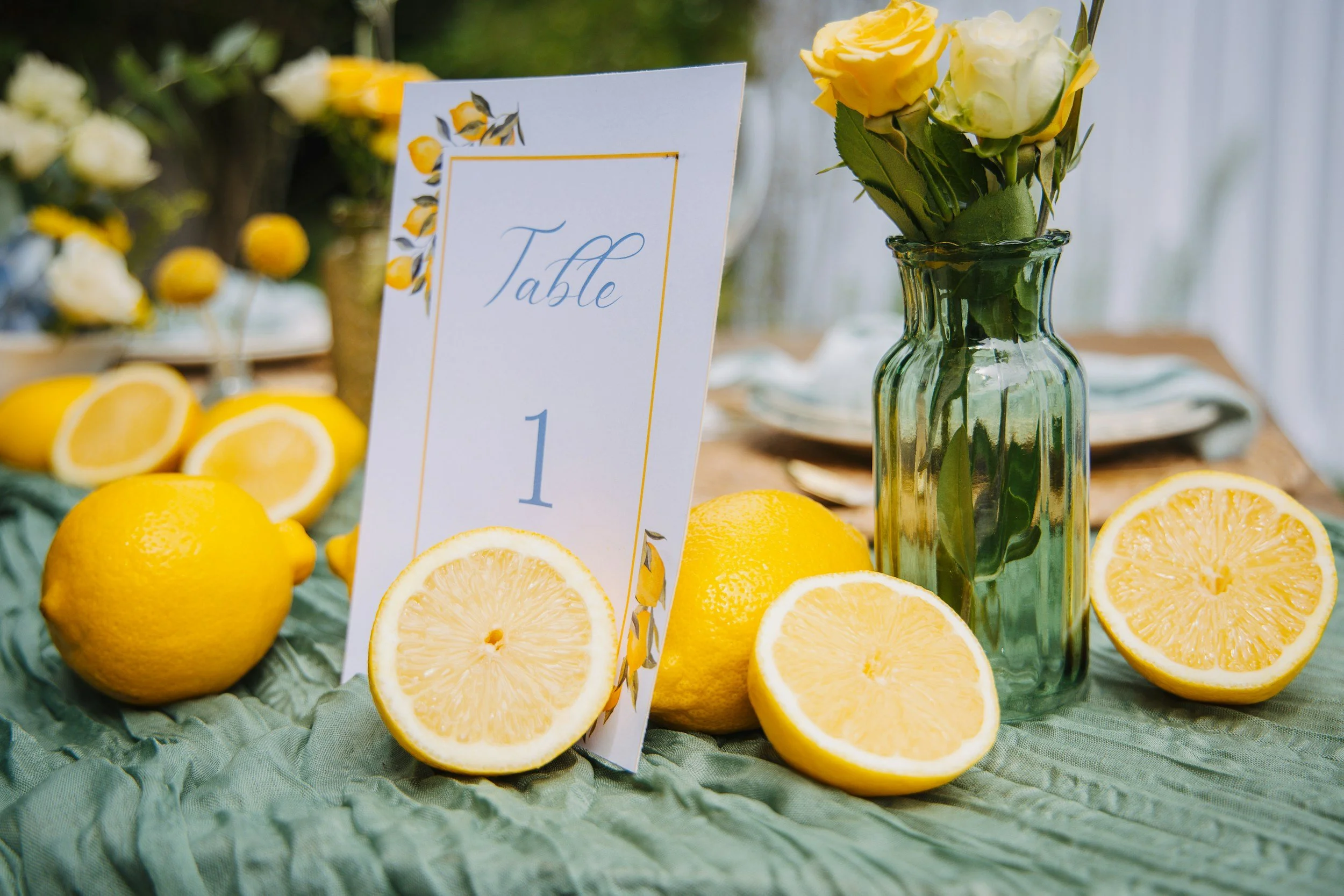 Table decorated with fresh lemons, lemon halves, a green glass vase with white and yellow roses, and a table number sign reading 'Table 1' in elegant script. The tablecloth is light green, and other lemon-themed decorations are visible in the background.