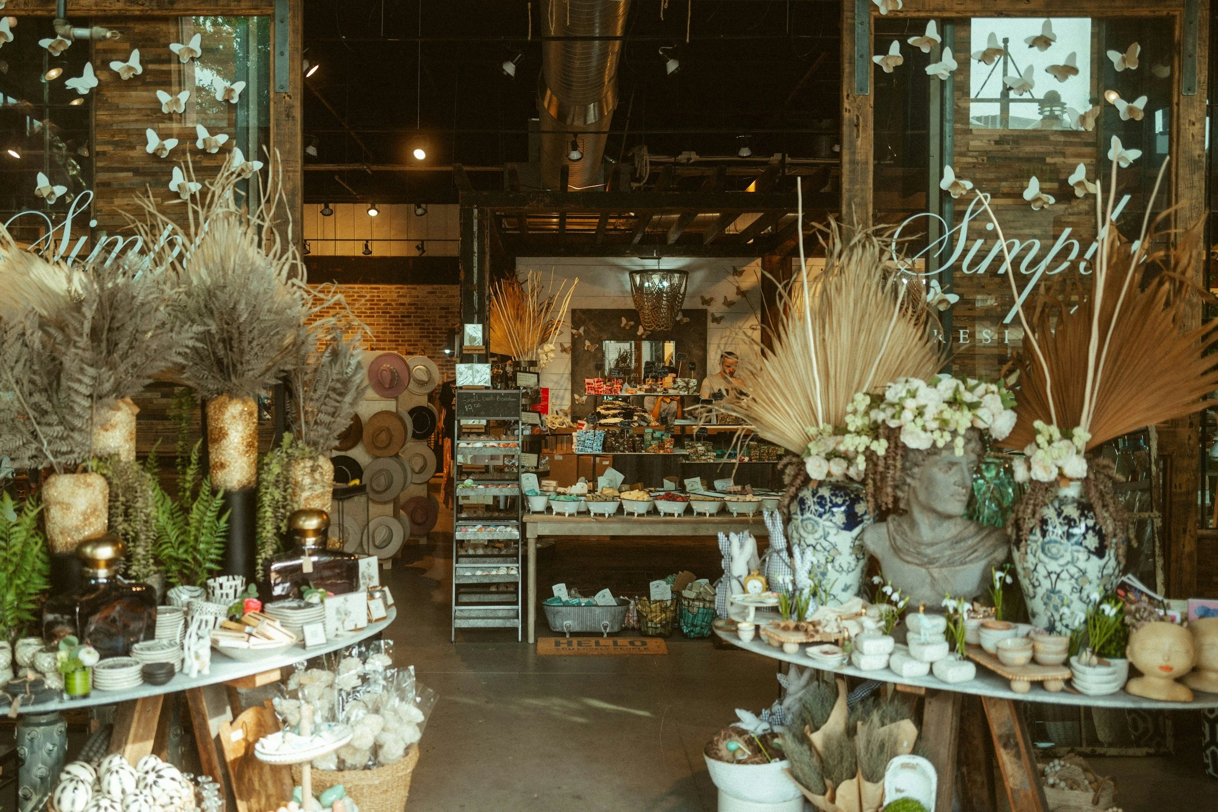 Inside a boutique store with shelves of hats, plants, vases, and decorative items, including large floral arrangements and butterfly wall art.
