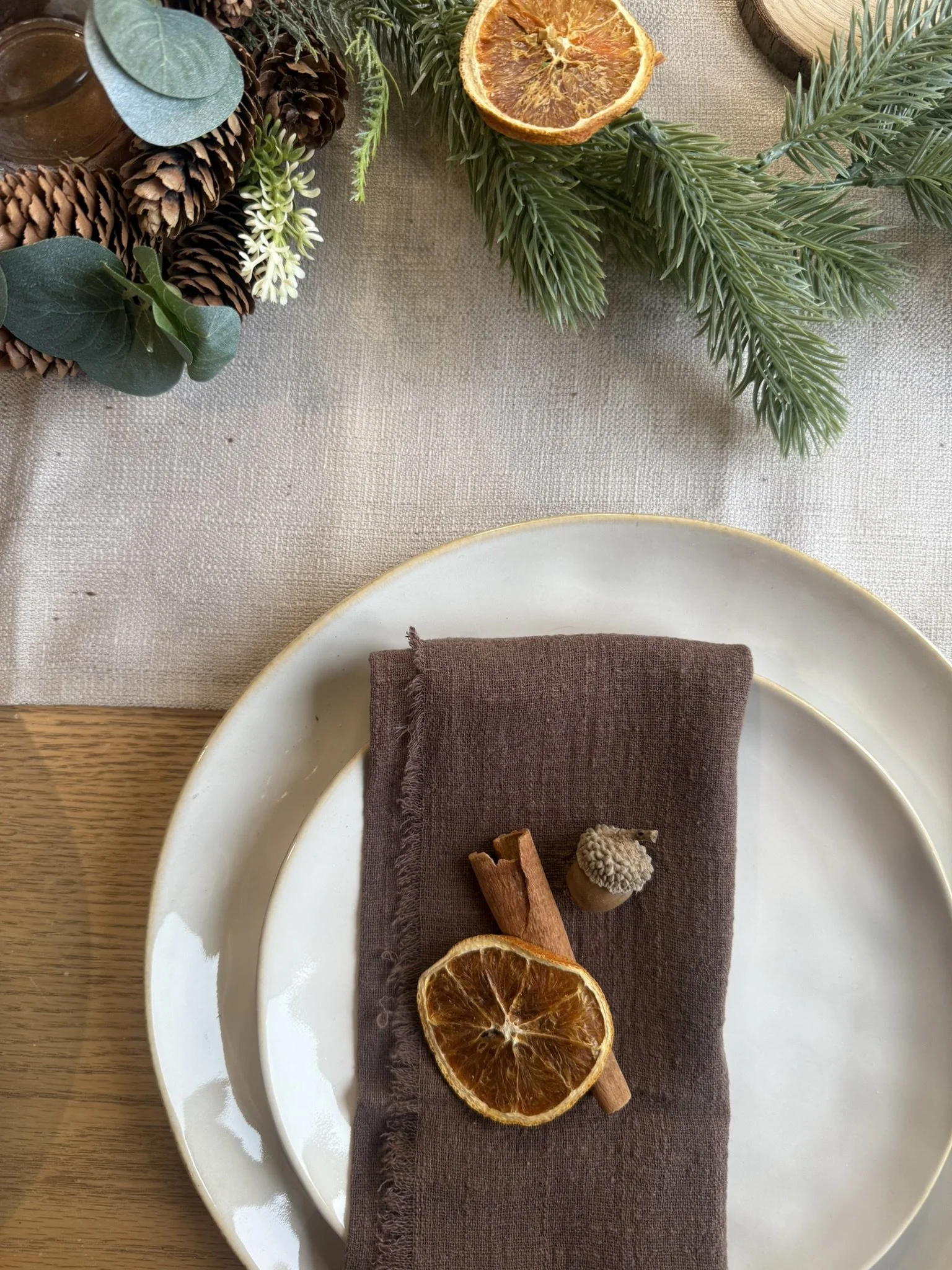 Table setting with a white plate, a dark brown napkin, a dried orange slice, a cinnamon stick, and a small pine cone, with Christmas greenery and dried orange slices on a linen tablecloth.