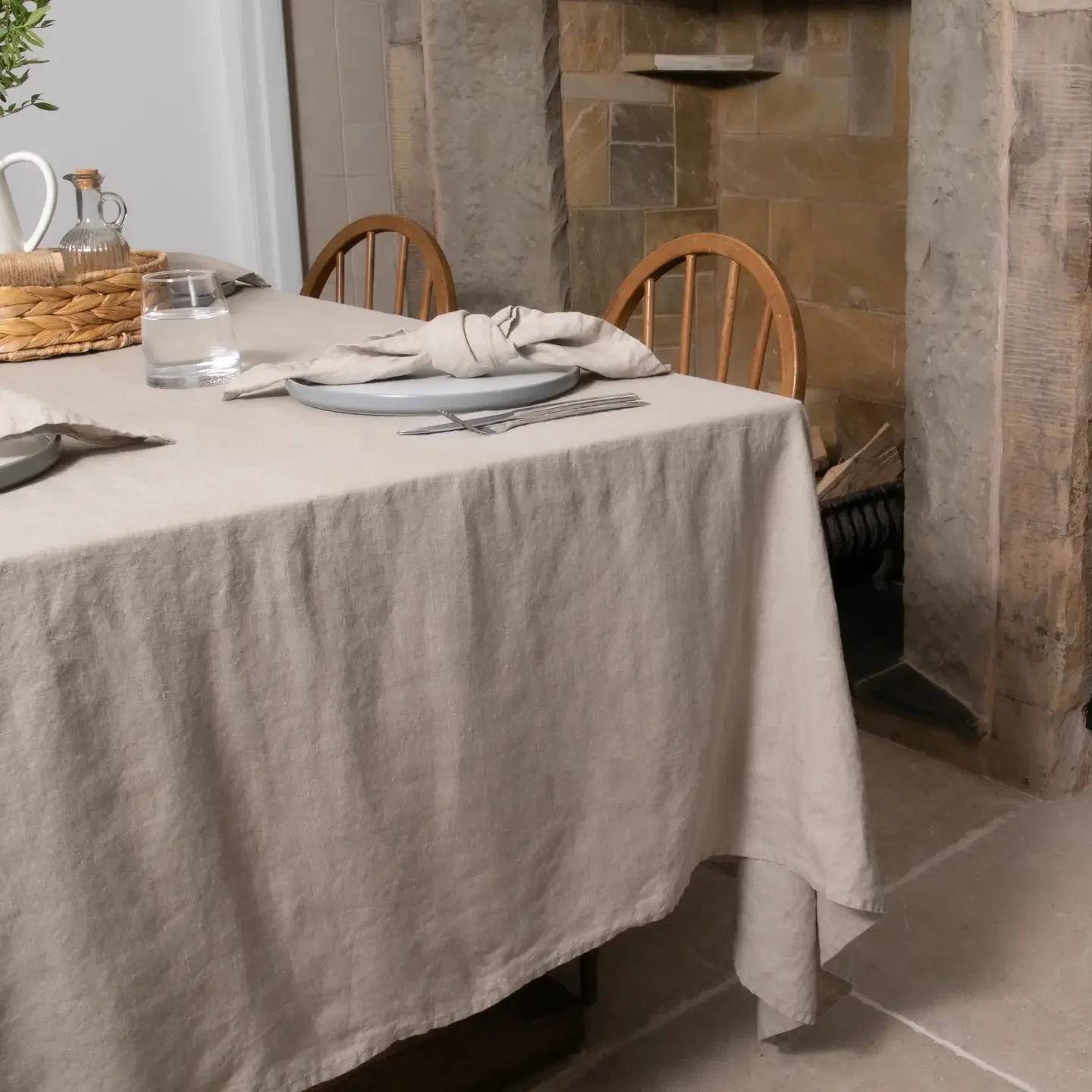 A dining table with a beige linen tablecloth, set with a gray plate, fork, knife, a cloth napkin, a glass of water, and a small tray with a glass pitcher and a small container, in a rustic style room with stone walls and wooden chairs.
