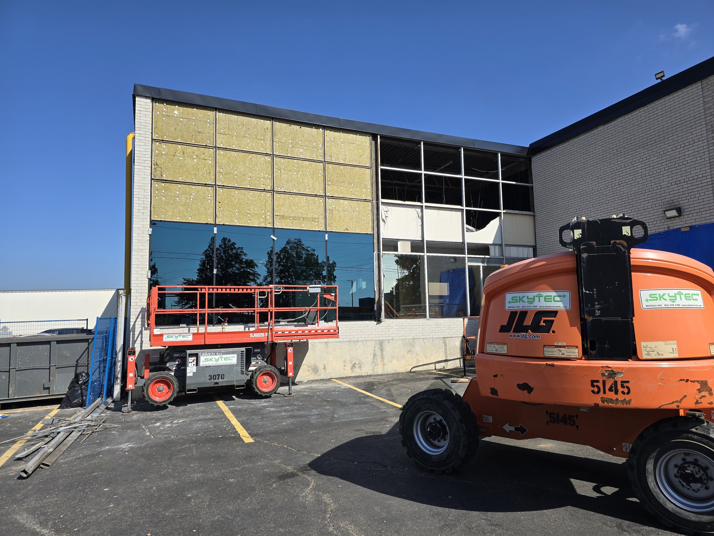 Construction site with a building under renovation, featuring a partially removed exterior wall, construction lift, and equipment in a parking lot under a clear blue sky.