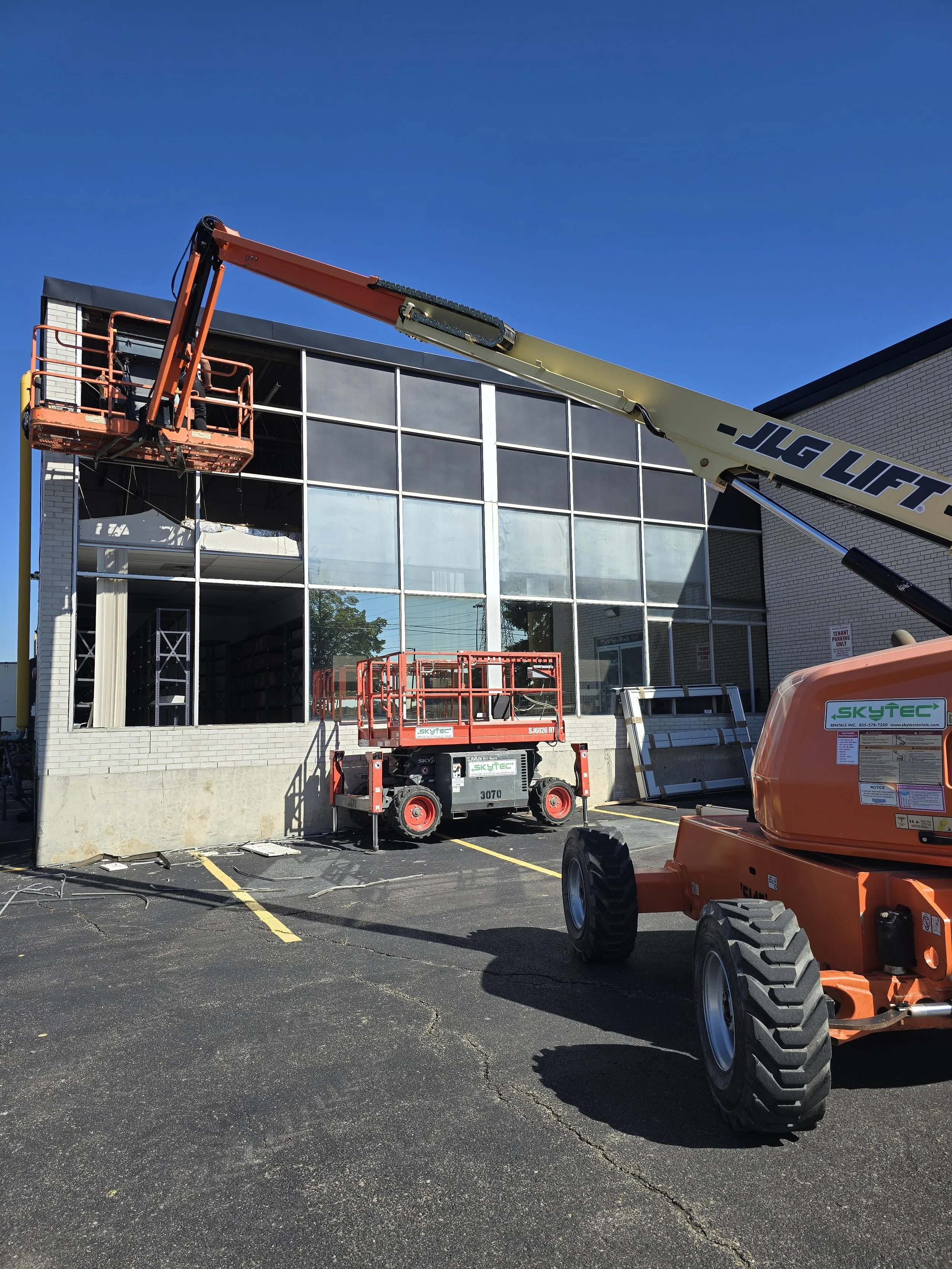 Construction workers using a boom lift to upgrade the exterior of a glass-front building under clear blue sky.