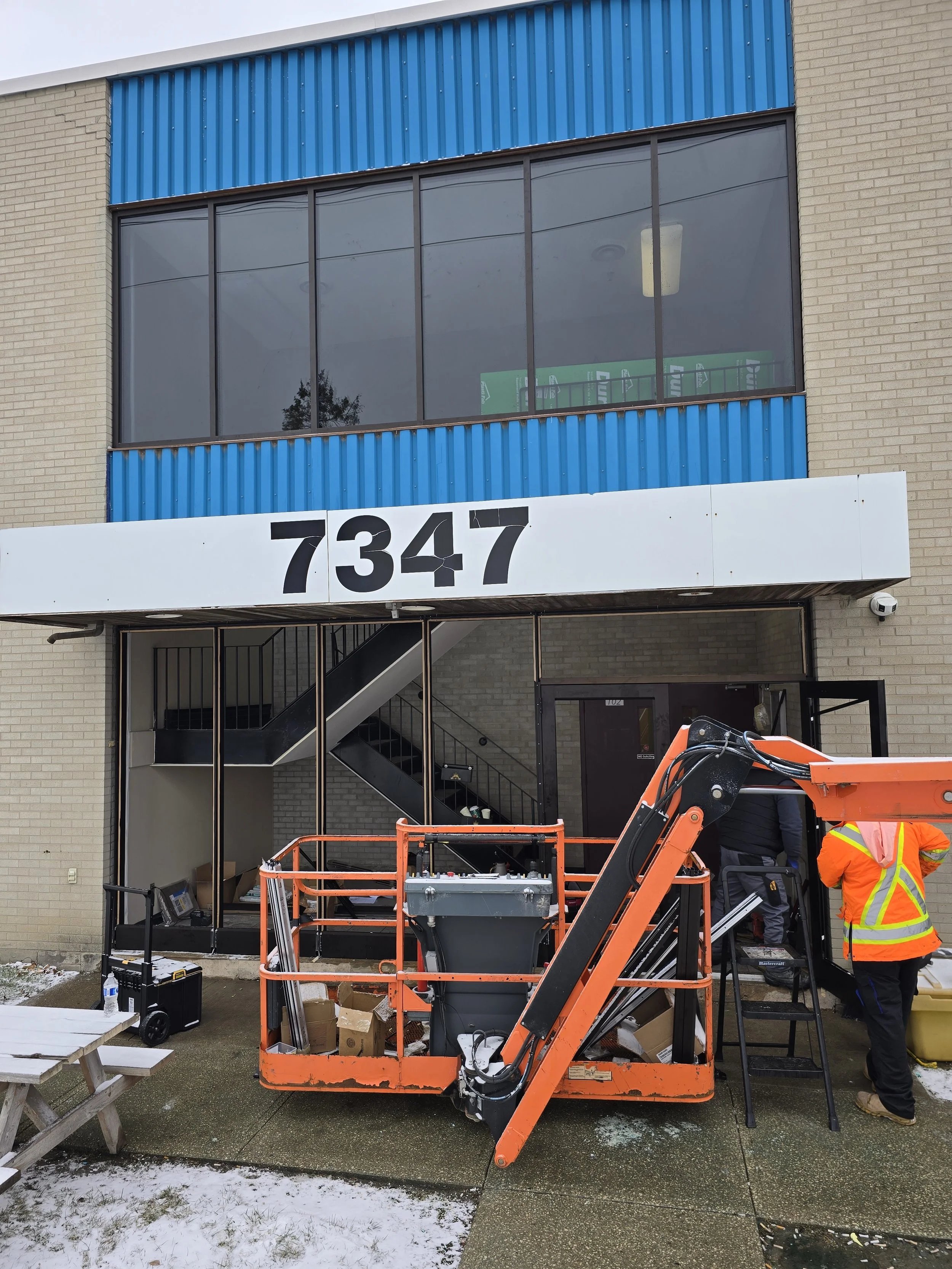 Construction workers installing a new entrance door at a commercial building with the address 7347 displayed above. The building has beige brick walls, large windows, and blue siding accents. There is construction equipment and materials in front, with some snow on the ground.