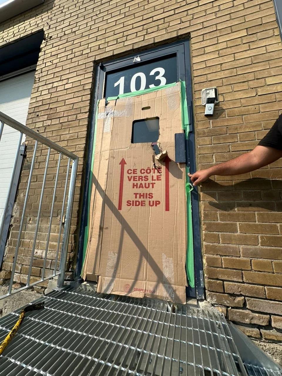 A brick building with a door covered by a large cardboard box with red text and arrows, indicating the side up. A person is holding the edge of the box, and the door has a sign reading '103'. There is a metal grate outside the door and a railing on the side.