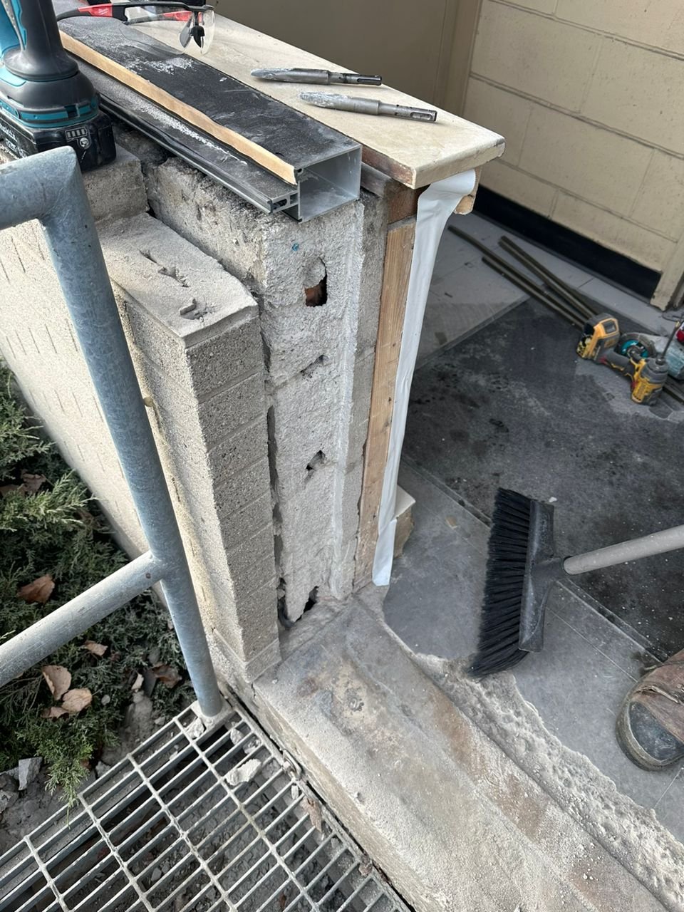 Construction site showing a concrete block wall, a workbench with tools, and cleaning tools like a broom, with construction materials and equipment nearby.