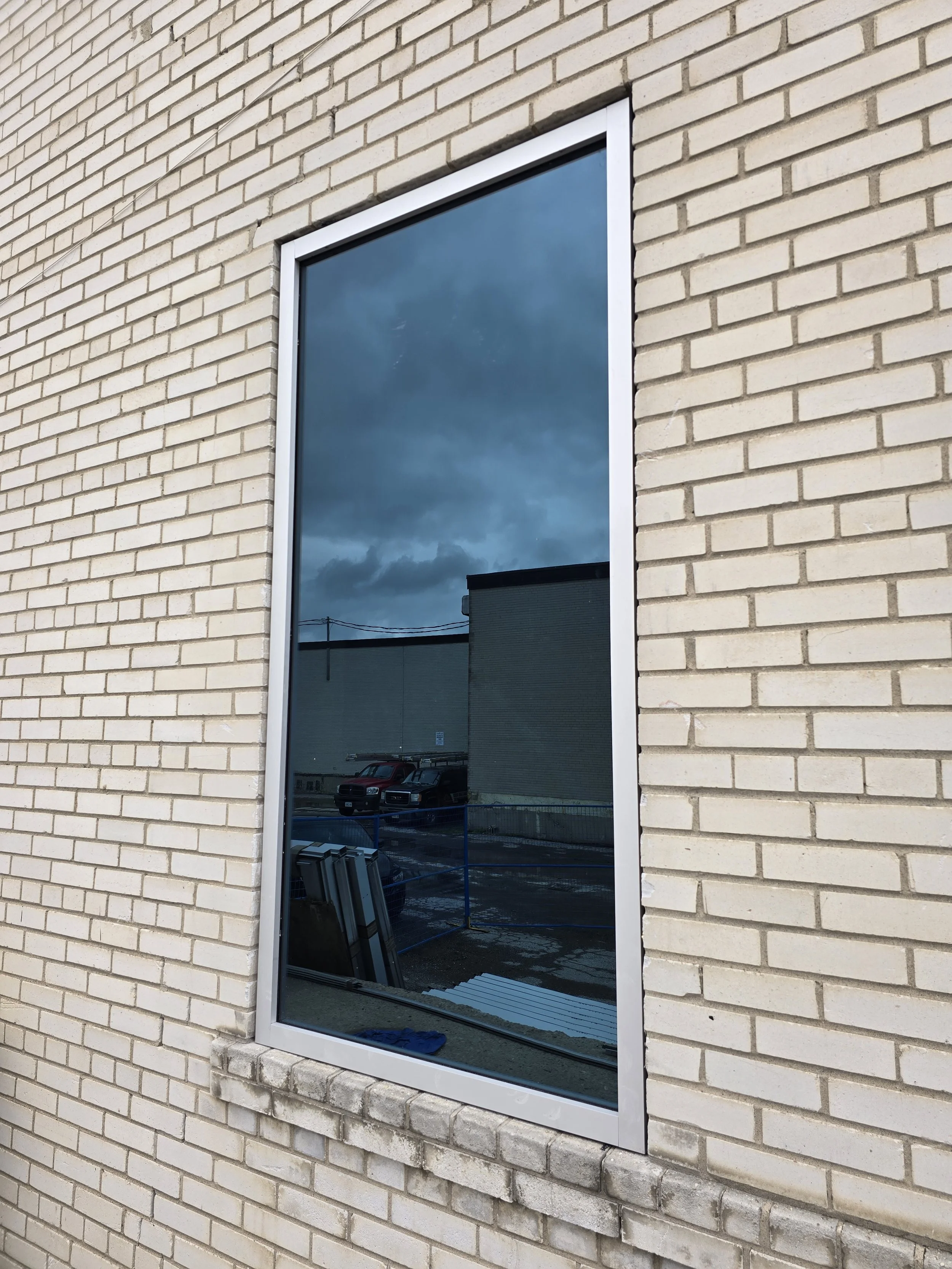 A reflective window on a beige brick building, showing a cloudy sky, parking lot, parked cars, and nearby buildings.
