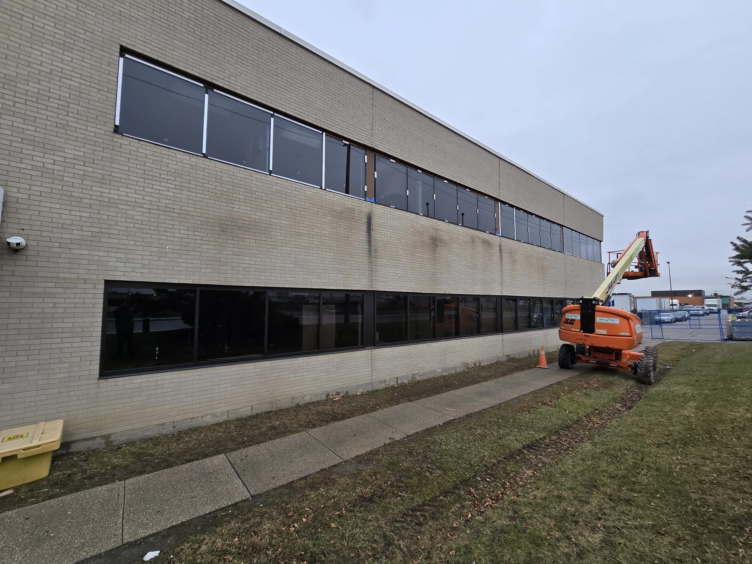 Construction worker using a cherry picker lift near the second story windows of a brick building, with a safety cone and the sidewalk in the foreground.