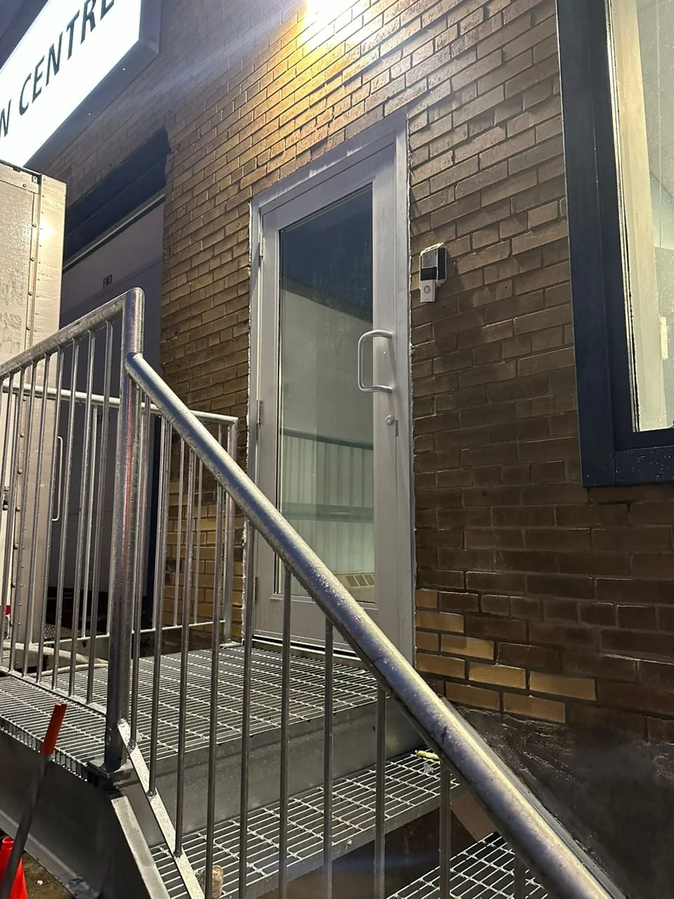 Exterior view of a building with a glass door, brick wall, metal ramp, handrails, and a security camera mounted on the wall.